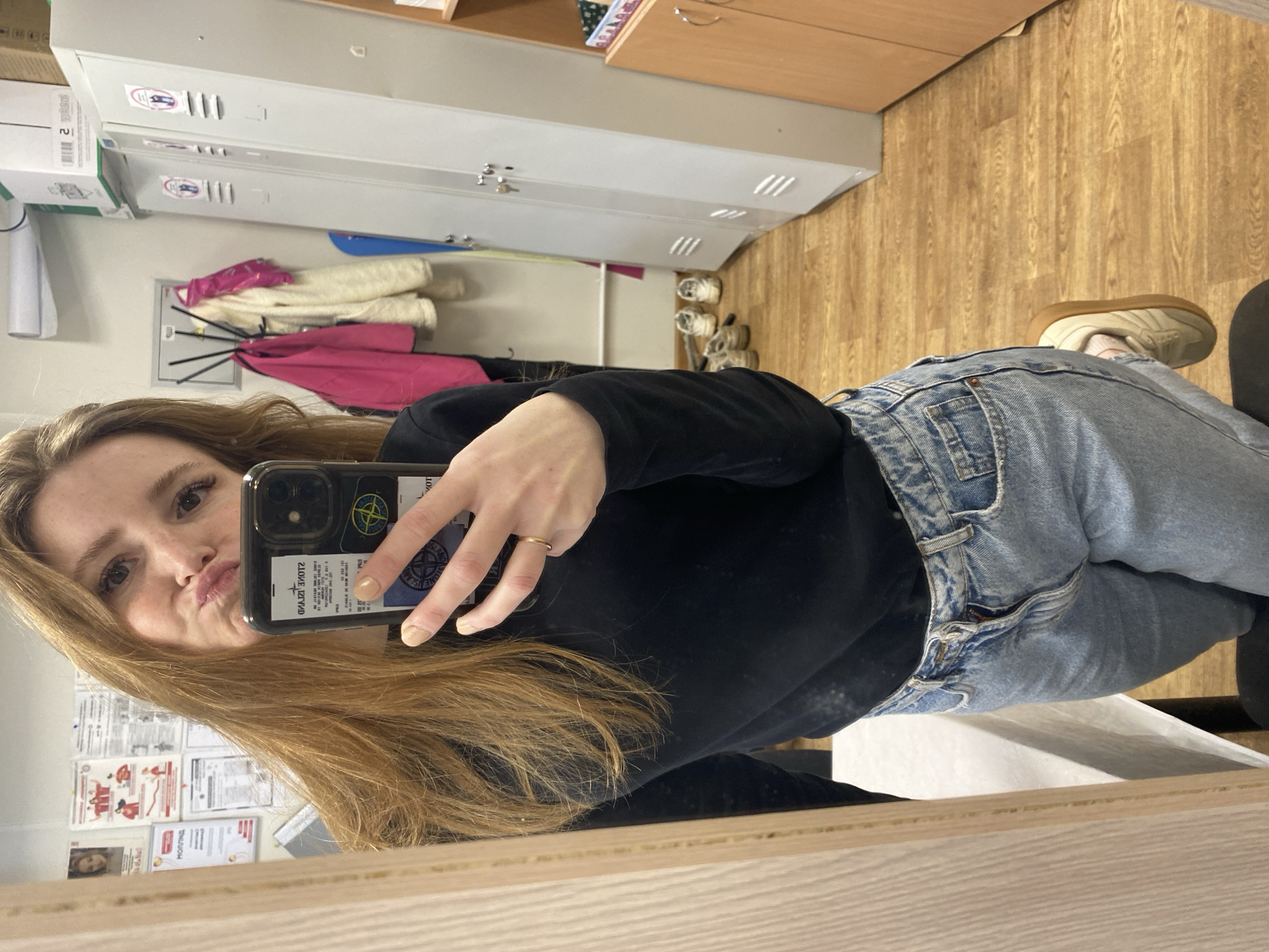 A young woman takes a mirror selfie in what appears to be an indoor common area or changing room, possibly in Chaikovsky, Russia. The image captures her from a slightly low angle, showing her upper body and legs. She has long, light brown hair and is looking directly into the phone held in her right hand, making a playful 'duck face' expression. She is wearing a black long-sleeved top, light blue denim jeans, and light-colored sneakers with a gum sole. A simple gold-colored ring is visible on her right ring finger, and her nails have light-colored polish.
The phone, likely an iPhone, is encased in a clear cover adorned with several stickers. Most notably, a prominent circular 'Stone Island' logo (compass rose design) is visible, along with some reversed text on another sticker, including 'STONE ISLAND'.
The background, a reflection in the mirror, reveals a functional indoor setting. On the left, several light-colored lockers or storage units are stacked, with white boxes placed on top, one clearly marked with the number '2'. A coat rack below the lockers holds a bright pink jacket, a cream-colored fluffy jacket, and another dark garment. Several pairs of shoes are placed on the light-colored wooden floor near the rack. The wall behind features various reflected documents, posters, and a small picture of a person; some faint Cyrillic text is visible but largely unreadable. The overall scene suggests a casual moment in an everyday interior environment.