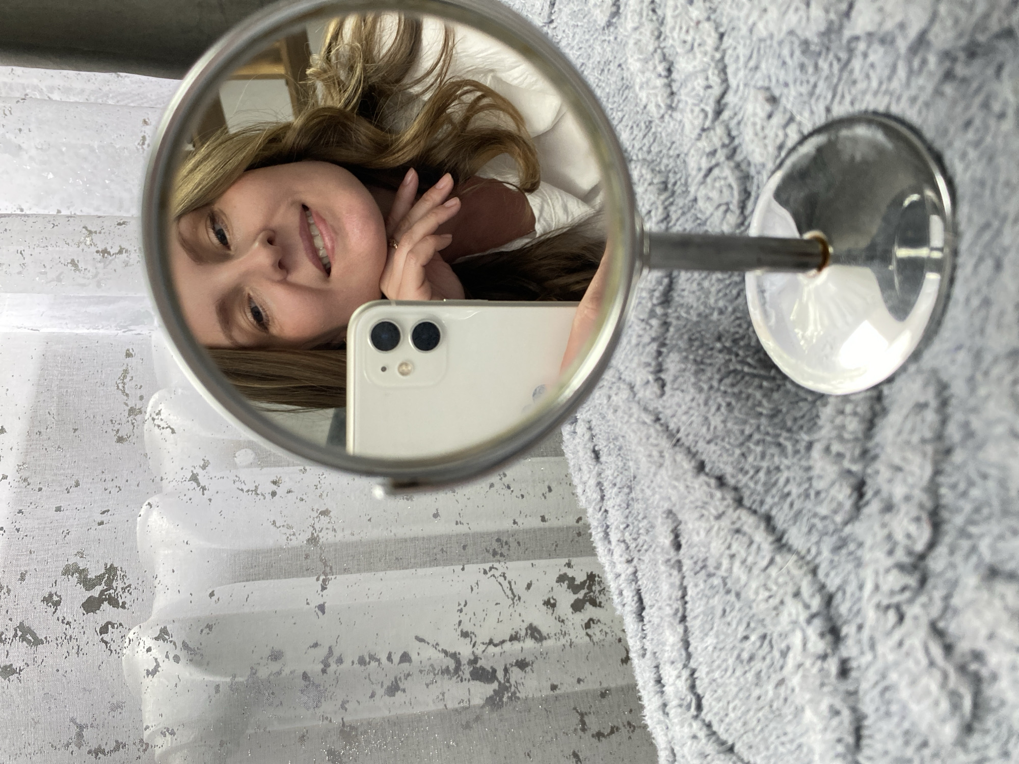 A woman with light brown hair is reflected in a round mirror. She is smiling and holding a white smartphone. The mirror has a metal stand. The background includes white patterned curtains and a gray textured surface, possibly a blanket or rug. There is no indication in the image that the photograph was taken in Noviy, Russia.