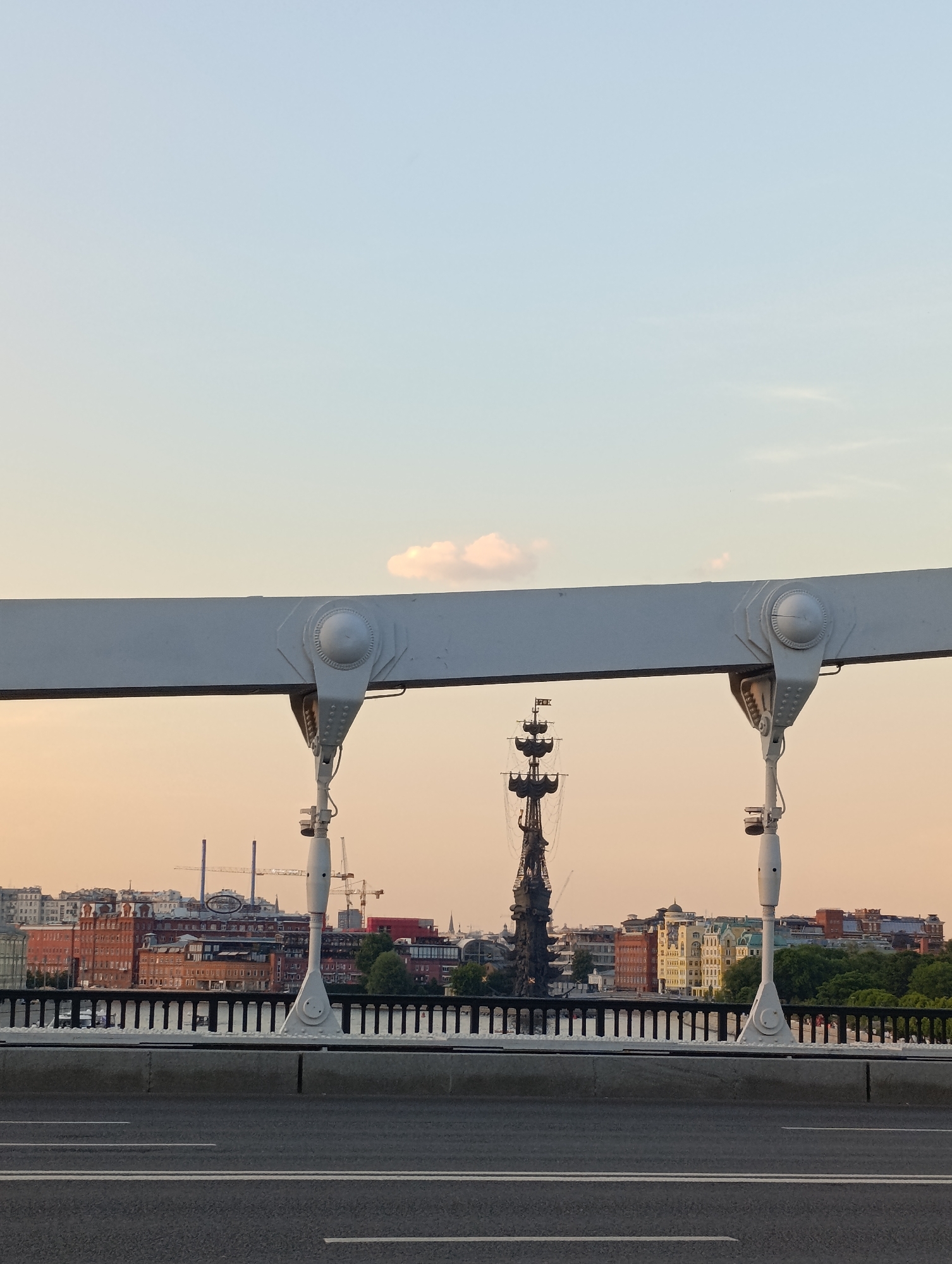 This image captures a panoramic view of the Moskva River and parts of Moscow's cityscape, primarily dominated by the Peter the Great Statue, seen from a bridge at dusk or dawn.
In the immediate foreground, an asphalt road surface with double white solid lines is visible. Above the road, a low concrete barrier separates it from the main bridge structure. Rising from this barrier are white, stylized vertical suspension elements of the bridge, which are wide and flat at the top, supported by conical structures with prominent circular bolt caps and riveted connections. These elements frame the central view of the distant cityscape. Between these bridge supports, a black metal railing with vertical balusters is visible.
Beyond the railing, the calm surface of the Moskva River is visible. Across the river, the dark silhouette of the Peter the Great Statue stands prominently against the horizon, its ship-like base and tall masts detailed even in shadow. The statue is located at the confluence of the Moskva River and the Vodootvodny Canal, specifically at the western tip of Bolotny Island.
To the left of the statue, a cluster of red-brick buildings, including one with two distinctive industrial chimneys (likely the former Red October chocolate factory complex), are visible along the riverbank. Further into the background, a mix of buildings with various architectural styles and colors—including yellow and light-colored facades—form the urban skyline. Several construction cranes are discernible on the distant horizon, indicating ongoing development.
The sky transitions from a pale blue at the top to a warm, soft peach and orange hue closer to the horizon, characteristic of twilight. A few small, scattered white clouds are present in the upper sky. The overall lighting is soft and diffused. No people or active interactions are clearly visible within the frame. The bridge in the foreground is identified as the Krymsky Bridge (Crimean Bridge), a suspension bridge in Moscow, Russia.