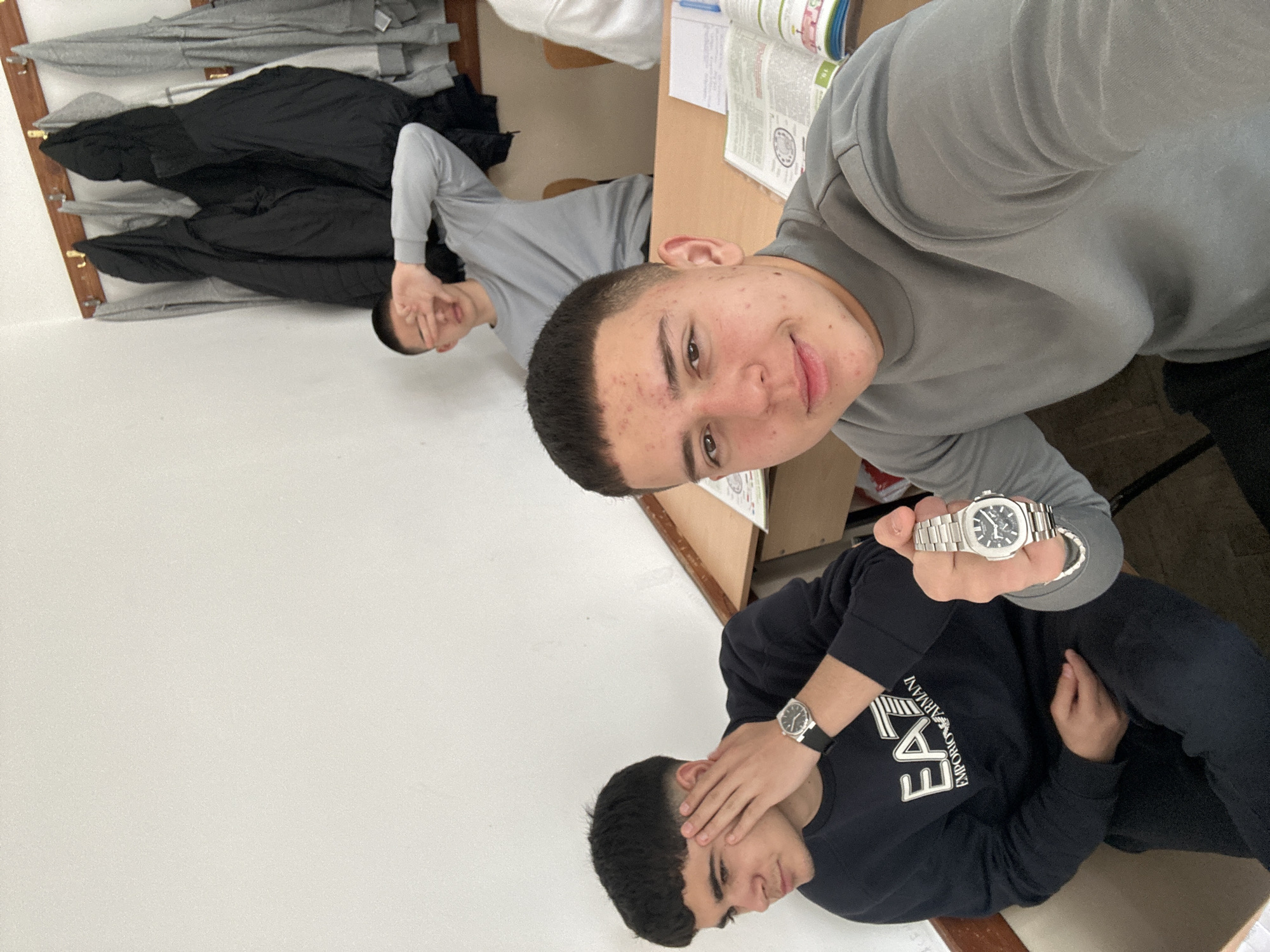 This image captures three young men indoors, likely in a casual setting such as a classroom or common room in Zlatograd, Bulgaria. The photo is taken from a tilted angle, focusing on the subjects.
In the foreground, a young man with a short, faded haircut and a grey long-sleeved shirt smiles towards the camera, holding up a silver-toned wristwatch with a dark face. He is seated at a light brown wooden desk, on which an open textbook or notebook with visible text and diagrams is placed.
To his right, another young man, also with a short haircut, is seated. He wears a black long-sleeved sweater with the logo "EMPORIO ARMANI EA7" prominently displayed on the front. His hands are covering his eyes, and a simpler black-strapped wristwatch is visible on his left wrist.
In the background, to the left, a third young man, similarly dressed in a grey long-sleeved shirt, is partially visible, also covering his eyes with his hand. He appears to be seated on the floor or a low bench against a white wall. On the far left, several coats, some grey and some black, hang on wooden hooks. The room features white walls and a wooden floor. The overall mood appears casual and playful.