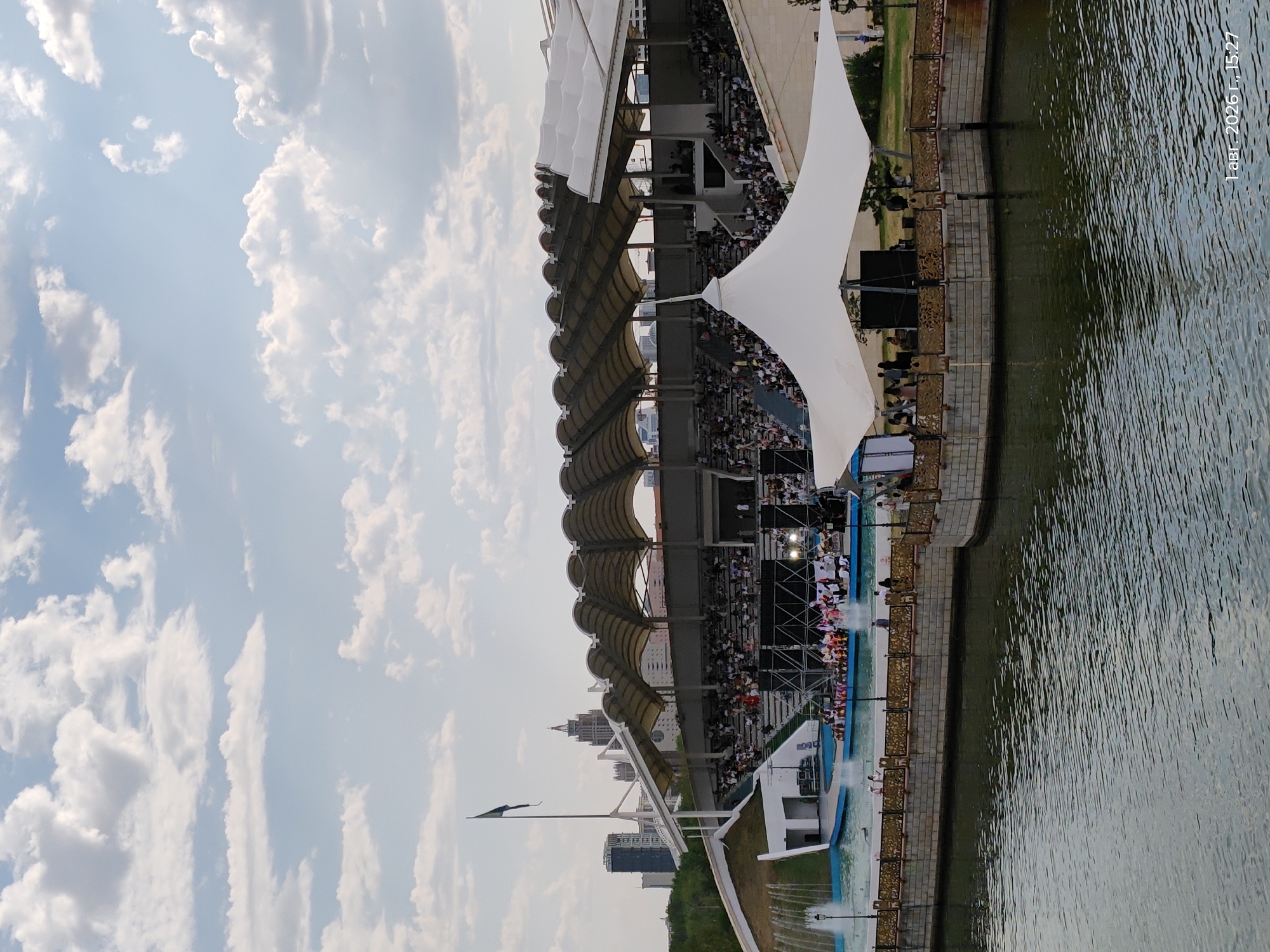 The image captures an outdoor performance area with a tiered seating section covered by a large, wavy roof structure, located in Astana, Kazakhstan. In the foreground, a body of water reflects the cloudy sky. To the left, a fountain sprays water into the air. A boat with performers in colorful attire is positioned in the water in front of the seating area. A white tent, possibly a stage canopy, is prominent on the right side of the image, set up on a stone platform overlooking the water. People are seated in the stands, facing the performance area. A flag is mounted on a tall pole to the left of the seating area, with modern city buildings visible in the background. The date and time stamp "1 авг. 2026 г., 15:27" is present in the lower right corner.