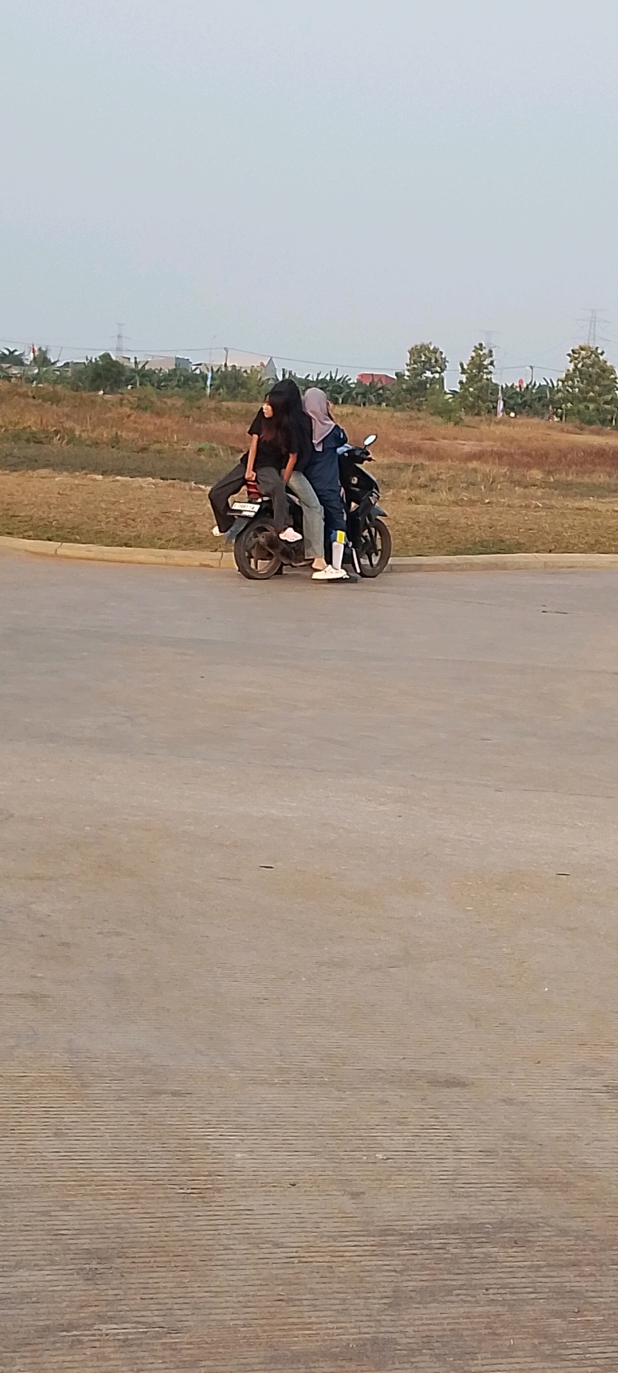 In Satriamekar, Indonesia, three individuals are seated on a black motorcycle on an asphalt surface. Two people are positioned in the front, with one person, wearing a dark hijab, appearing to be steering. Next to them, another person, also in dark clothing, is seated. A third person, in dark clothing, is seated behind them on the motorcycle. The background consists of a grassy field with scattered trees and some buildings in the distance, under a pale, hazy sky.