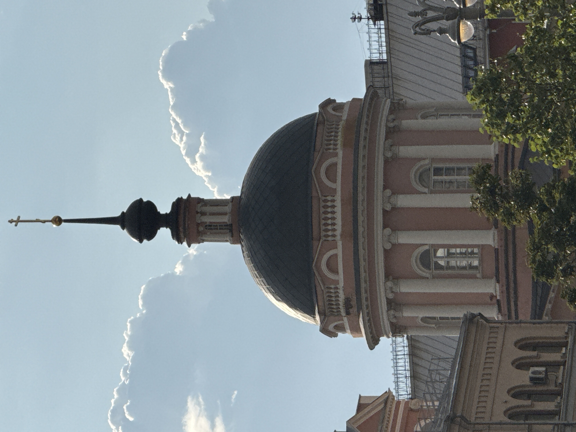 The image captures a close-up view of the pink rotunda dome and spire of the Church of the Ascension of the Lord in Nikitniki, Moscow, Russia, against a backdrop of fluffy white clouds and a clear blue sky. The dome is dark gray and topped with a slender, dark spire and a golden cross. The rotunda features white Corinthian columns and arched windows with white frames. Below the dome, the building's lower sections, also pink, are partially visible. To the left, a section of a cream-colored building with arched windows and an air conditioning unit is present. In the foreground and right side, lush green trees partially obscure the lower parts of the church and surrounding structures. A lamppost with a glowing orb is visible on the far right. The overall scene suggests a bright, sunny day.