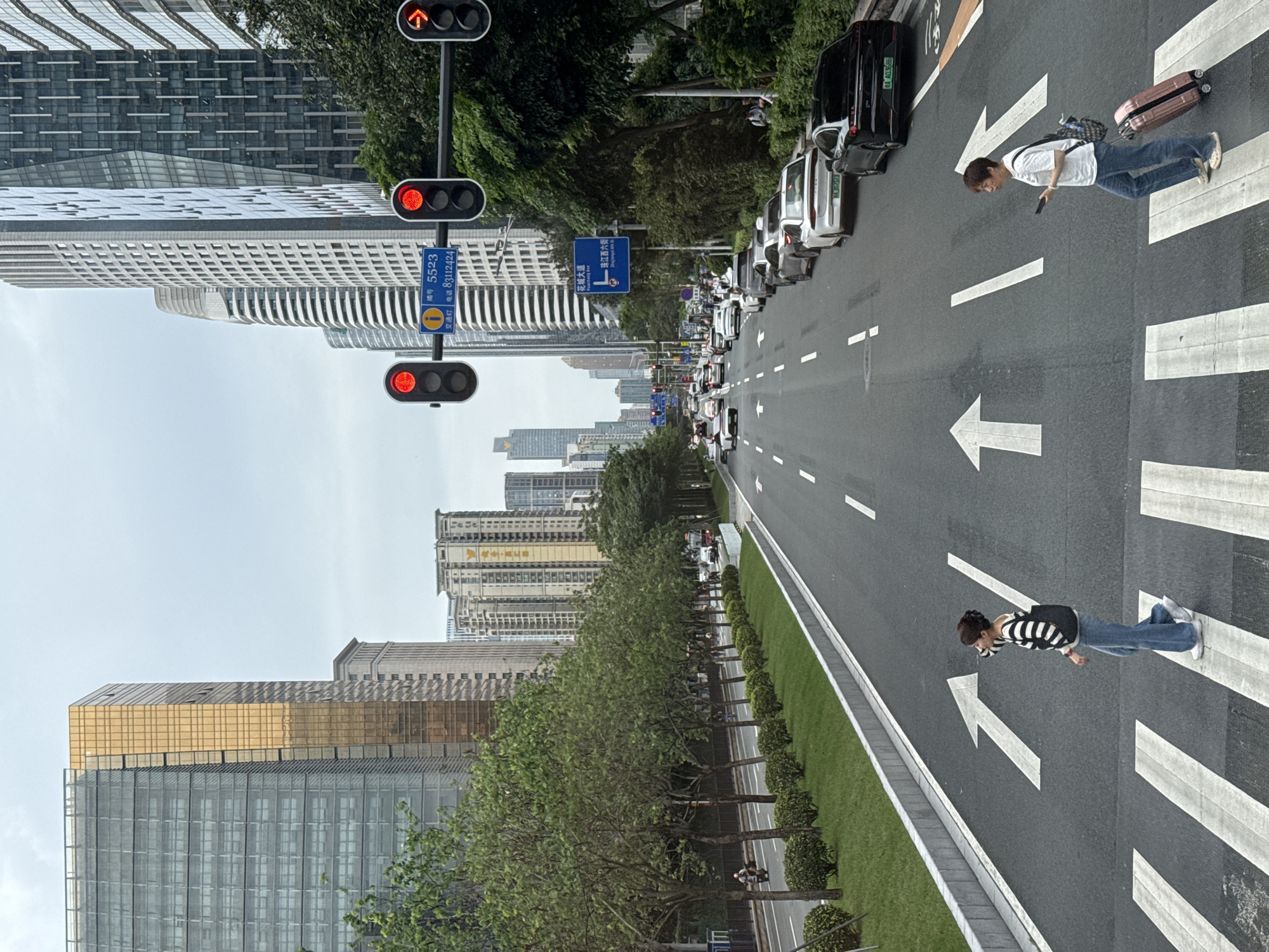 The image depicts a street scene in Tianhe District, China. A wide, multi-lane road is visible, with traffic stopped at a red traffic light. Two pedestrians are crossing the street in the crosswalk. One is a man wearing a white shirt and jeans, pulling a pink suitcase. The other is a woman with a striped shirt and jeans. The road is marked with white directional arrows. Green trees line the side of the road. Tall buildings are visible on either side of the street, including a gold-colored building on the left. A street sign in Chinese is visible near the traffic lights.