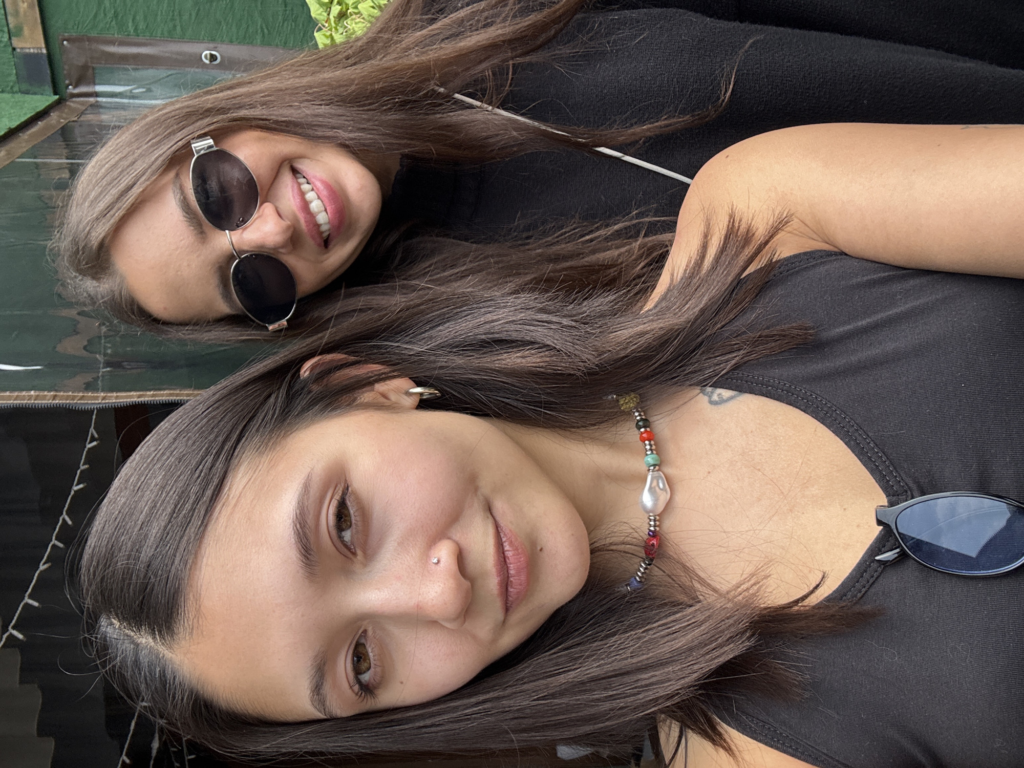 Two women are posing for a photo. The woman in the foreground has brown hair, brown eyes, and a nose piercing. She is wearing a black tank top and a beaded necklace. The woman in the background has long, brown hair and is wearing black sunglasses and a black top. The location is not identifiable as Vladivostok, Russia, based on the provided image. The environment appears to be outdoors, with a green surface visible in the background.