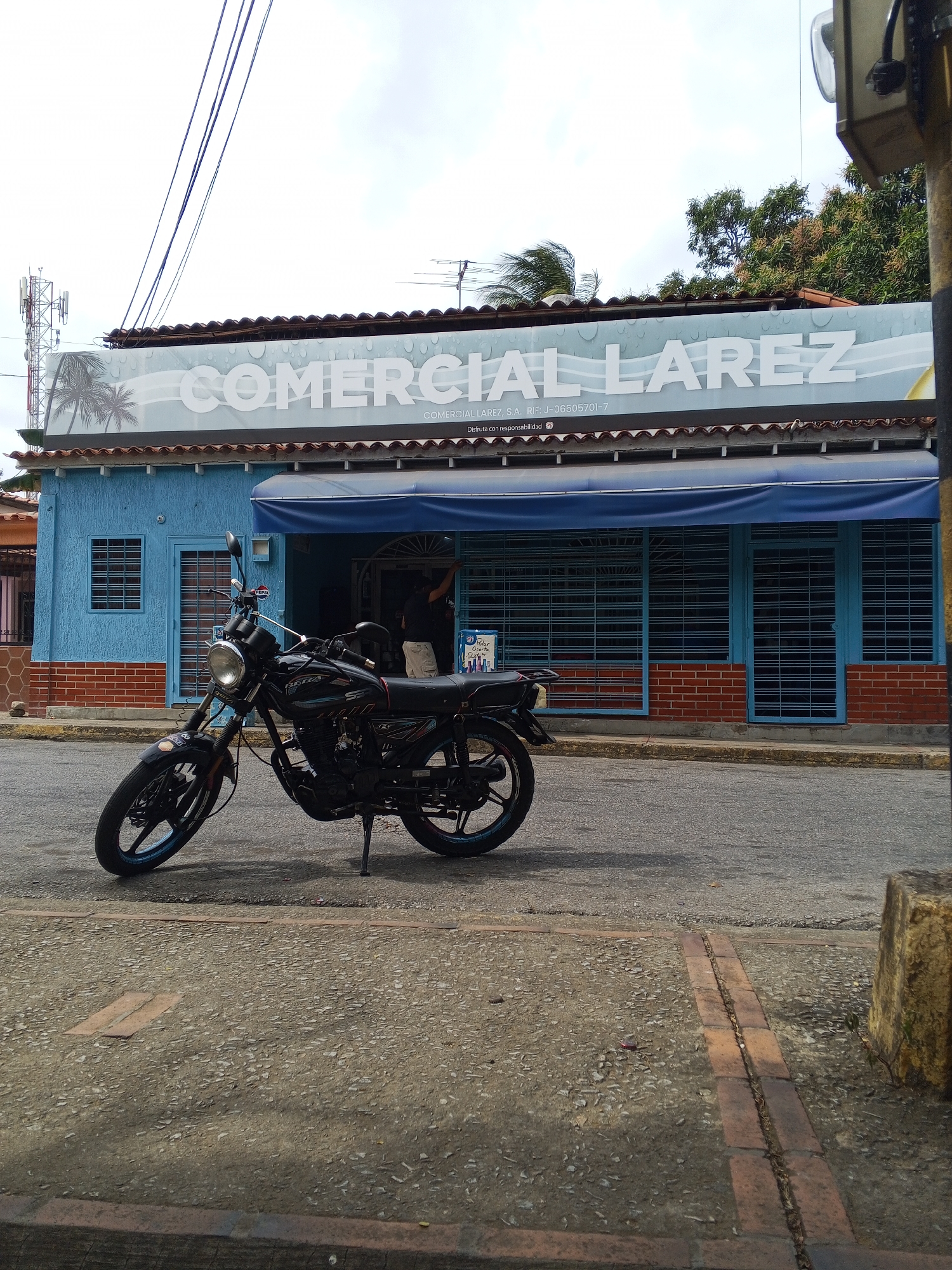 Here's a detailed description of the image:
**Setting:** The image appears to be taken on a street in front of a commercial building. The location is likely a city or town, with paved streets and a mix of residential and commercial properties.
**Building:** The building is painted blue with brick accents. A sign above the building reads "COMERCIAL LAREZ", also indicating it's the name of the establishment. The sign also displays "COMERCIAL LAREZ, S.A. RIF: J-06505701-7" and "Disfruta con responsabilidad" below it. There's a blue awning above the windows and doors. The windows and doors are secured with metal grilles, suggesting it might be closed or protecting against theft.
**Motorcycle:** A black motorcycle is parked on the street in front of the building. It is positioned at an angle, with the front wheel turned towards the building.
**Street and Sidewalk:** The street is paved, with a sidewalk visible in the foreground. The sidewalk is made of bricks.
**Environment:** The sky is overcast, suggesting it might be a cloudy day. Trees and other greenery are visible in the background.
**Other details:** A cellphone tower stands in the background, showing the presence of modern technology. A small box of soda cans can be seen at the store.