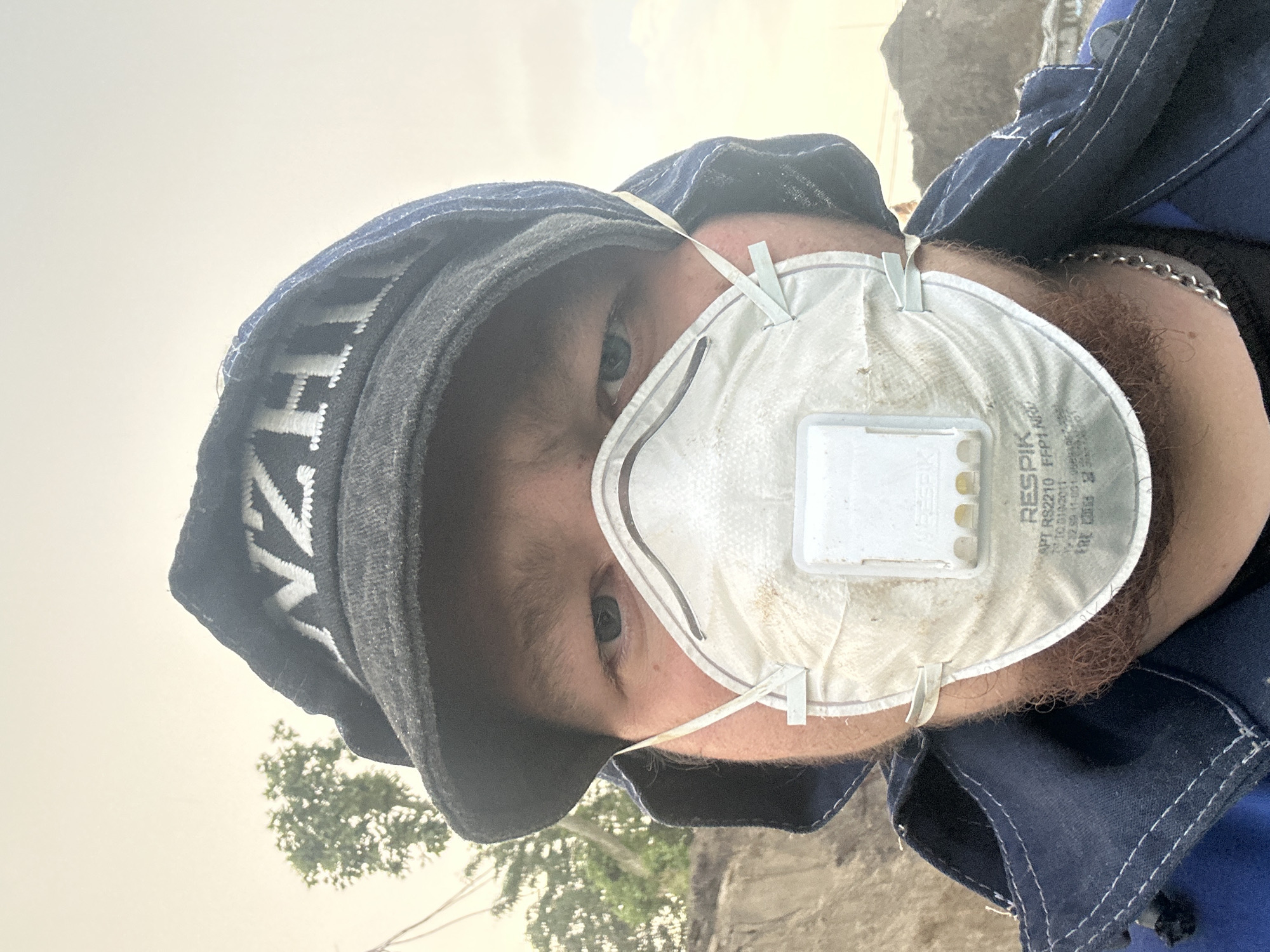 The image is a close-up, selfie-style photograph of a person wearing a dark-colored hooded jacket and a white respirator mask. The person also wears a dark beanie with white lettering that appears to read "NZH" or "NJH". The subject's eyes are visible above the mask, and they have light-colored irises. The background of the image shows an outdoor environment with a tree and what appears to be a dirt embankment or pile of material, possibly indicating a construction or outdoor work site. The sky is overcast. The image does not contain any specific landmarks or details that would definitively place it in Krasnodar, Russia.