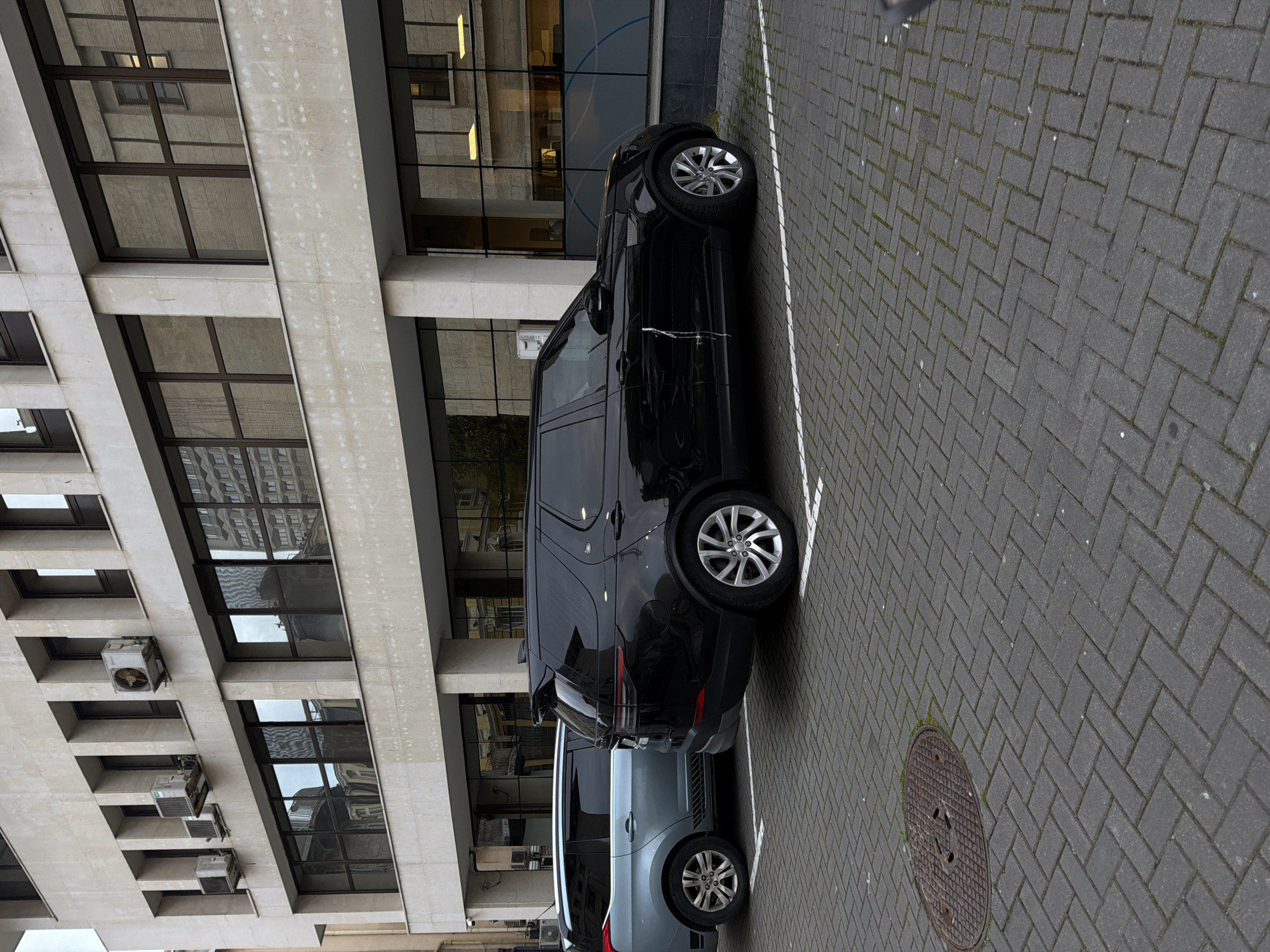 A black SUV is parked on a gray brick paved parking space, next to a building with multiple windows and several air conditioning units visible on the facade. Another blue/gray SUV is parked behind it. A manhole cover is visible near the parked cars. The image is rotated by 90 degrees clockwise. There is no information about the location in the city of Burgas, Bulgaria.