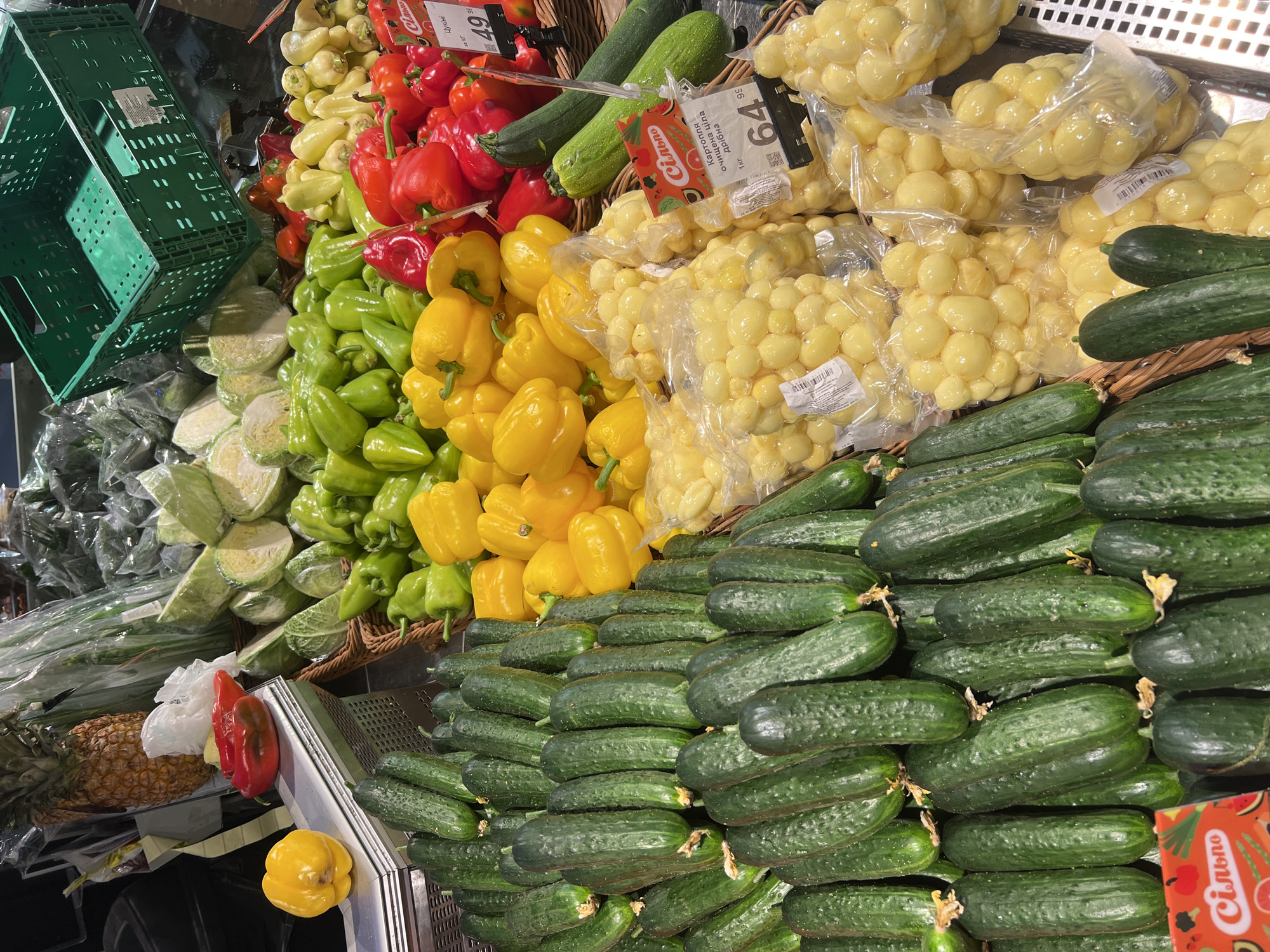The image shows a vibrant display of fresh produce at a grocery store, likely in Odesa, Ukraine, given the Cyrillic script on the price tags. In the foreground, a large pile of dark green cucumbers is neatly arranged in a basket. To the right of the cucumbers, several bags filled with small, yellow, round vegetables, possibly cherry tomatoes or small peppers, are displayed. Further back, a variety of colorful bell peppers are piled high, including bright red, yellow, and green varieties. Behind the peppers, there are bundles of green onions and a head of lettuce. In the upper left corner, a pineapple is visible. The overall impression is of a well-stocked produce section, with items attractively presented for sale. The lighting is bright, typical of a retail environment.