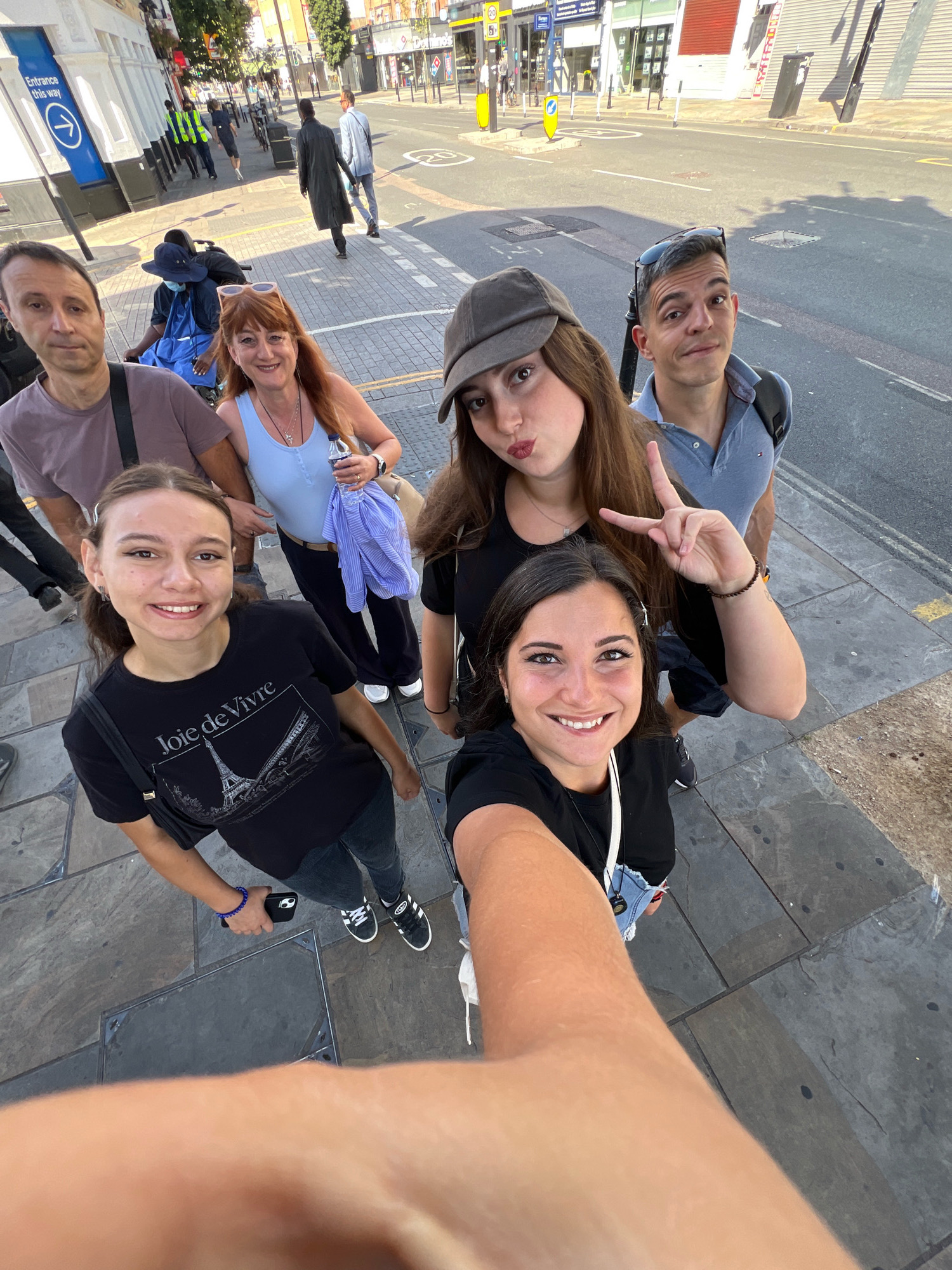 The image depicts a group of six individuals posing for a selfie on a paved sidewalk in front of various shops and a street. The group consists of three women and three men.
In the foreground, a woman in a black t-shirt with a graphic print of the Eiffel Tower is smiling at the camera, holding a phone with her right hand to take the selfie. To her left, another woman, also smiling, is visible from the shoulders up. She is wearing a light blue tank top and has a bottle of water in her hand.
Behind them, a man in a grey polo shirt is looking towards the camera with sunglasses on his head. To his left, a woman with long brown hair is making a peace sign with her right hand. She is wearing a dark grey baseball cap.
Further back, a man in a purple t-shirt is partially visible on the left side of the frame. Next to him, another man in a dark coat is walking away from the camera. In the background, several people are walking on the street and sidewalk. One person is in a wheelchair. The setting appears to be a commercial street in London, United Kingdom, as indicated by the architectural style of the buildings and the presence of a blue sign with white text that reads "Entrance this way." The street is paved with asphalt and marked with lane lines. The time of day appears to be daytime, with sunlight casting shadows on the ground.