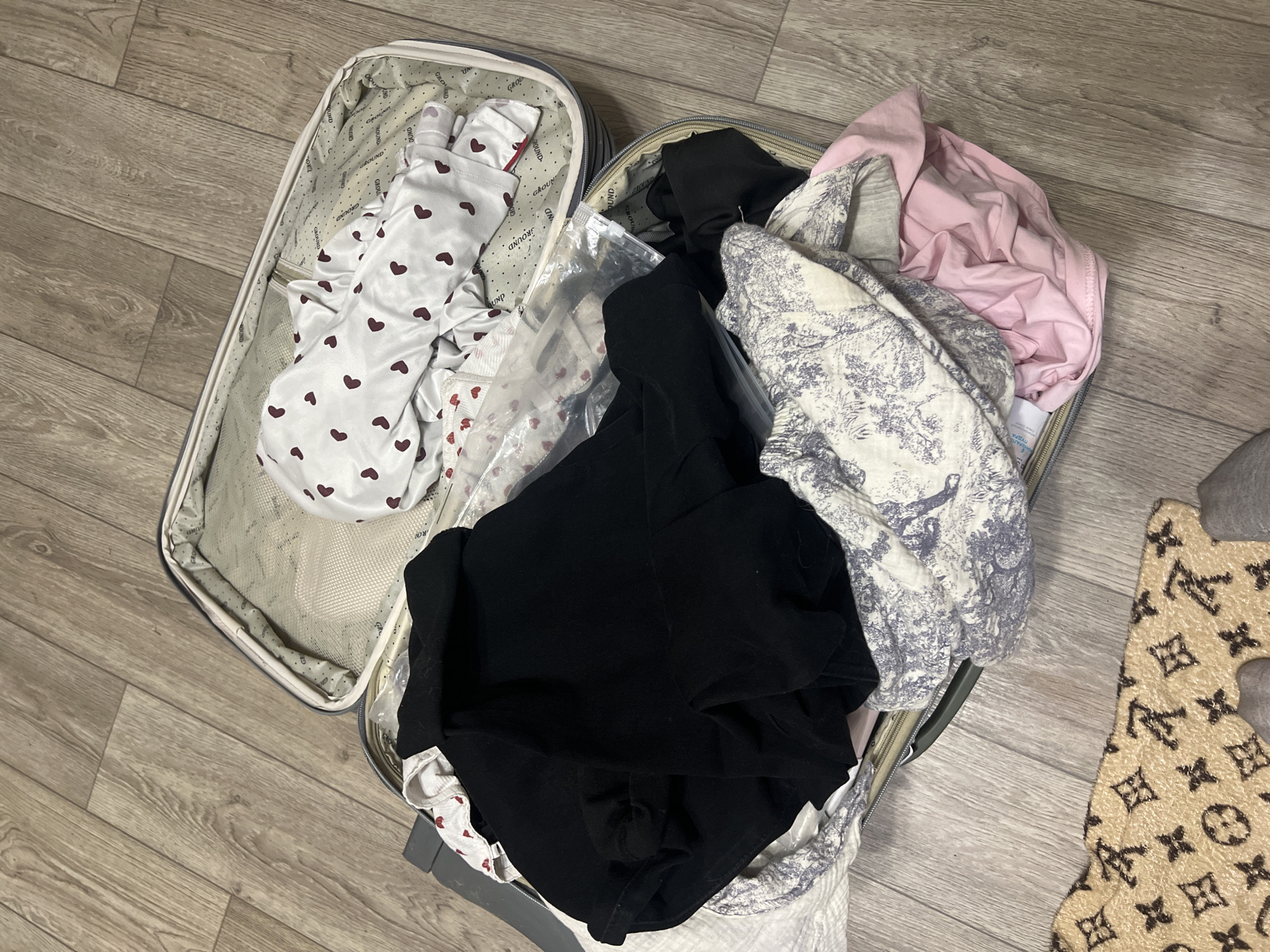 The image shows an open suitcase on a wooden floor. The suitcase is filled with various clothing items, including a white garment with red hearts, a black garment, a white and blue patterned garment, and a pink garment. A clear plastic bag is also visible among the clothing. The location is in Chaikovsky, Russia. A portion of a patterned rug is visible in the lower right corner.