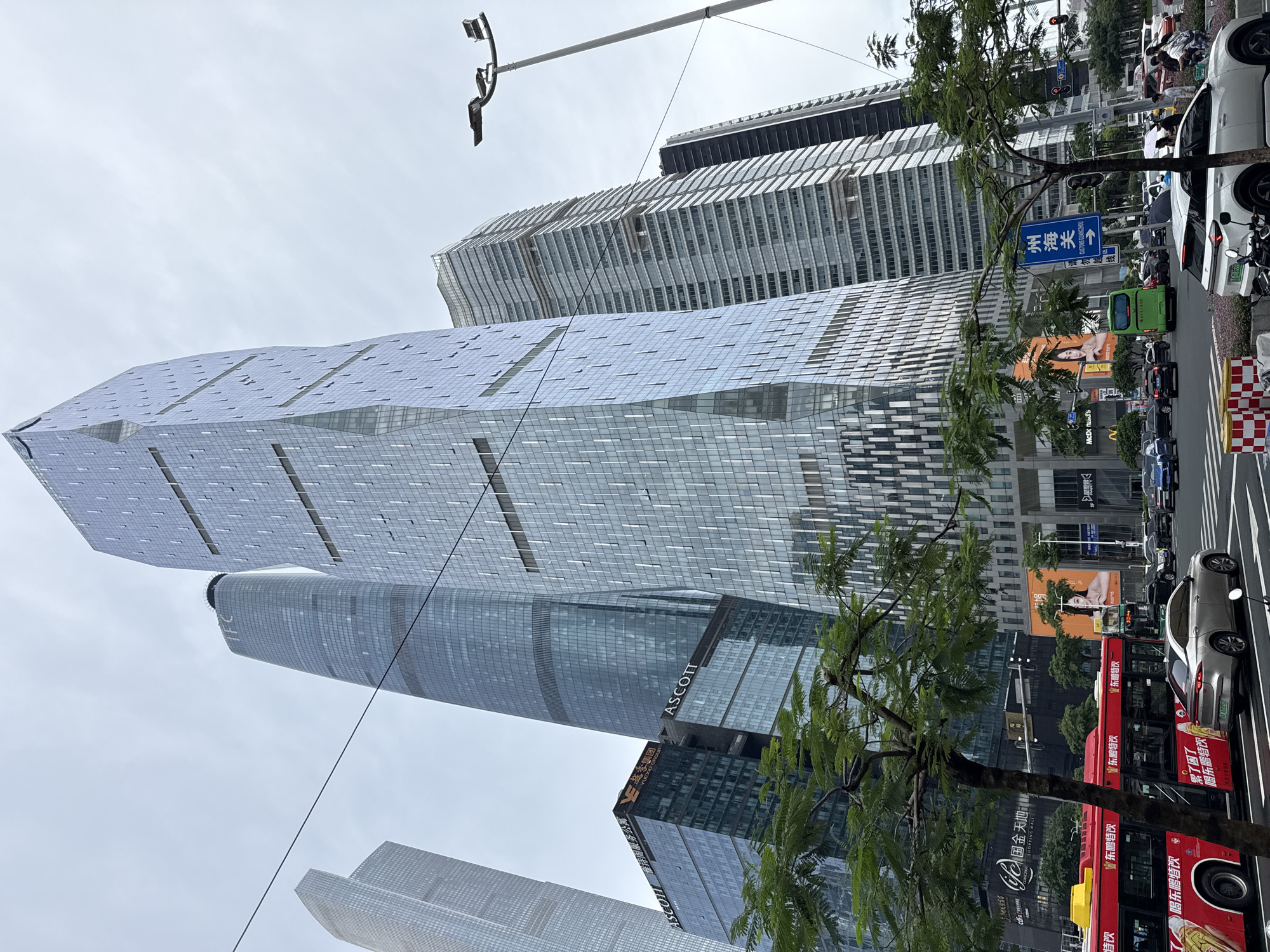 This is a wide-angle, eye-level, outdoor, daytime photograph taken in Tianhe District, China. The scene captures a modern urban environment dominated by several tall, glass-facade skyscrapers. The tallest building in the foreground has a distinctive, angular design with a shimmering, multi-faceted surface. To its left, a darker, cylindrical skyscraper with the "IFC" logo and a helipad is visible. Another skyscraper to the right features a layered, horizontal design. In the mid-ground, a lower building with prominent "ASCOTT" signage is visible.
The street level shows a busy intersection with traffic, including a red double-decker bus on the left, several cars, and a green bus. Pedestrians can be seen near the crosswalks. Large digital billboards are mounted on the buildings, displaying advertisements. Green trees with lush foliage line the street, partially obscuring some of the buildings. The sky is overcast, with a diffused, pale light. A streetlamp with multiple lights is positioned on the right side of the frame. The overall impression is one of a bustling commercial and financial district.