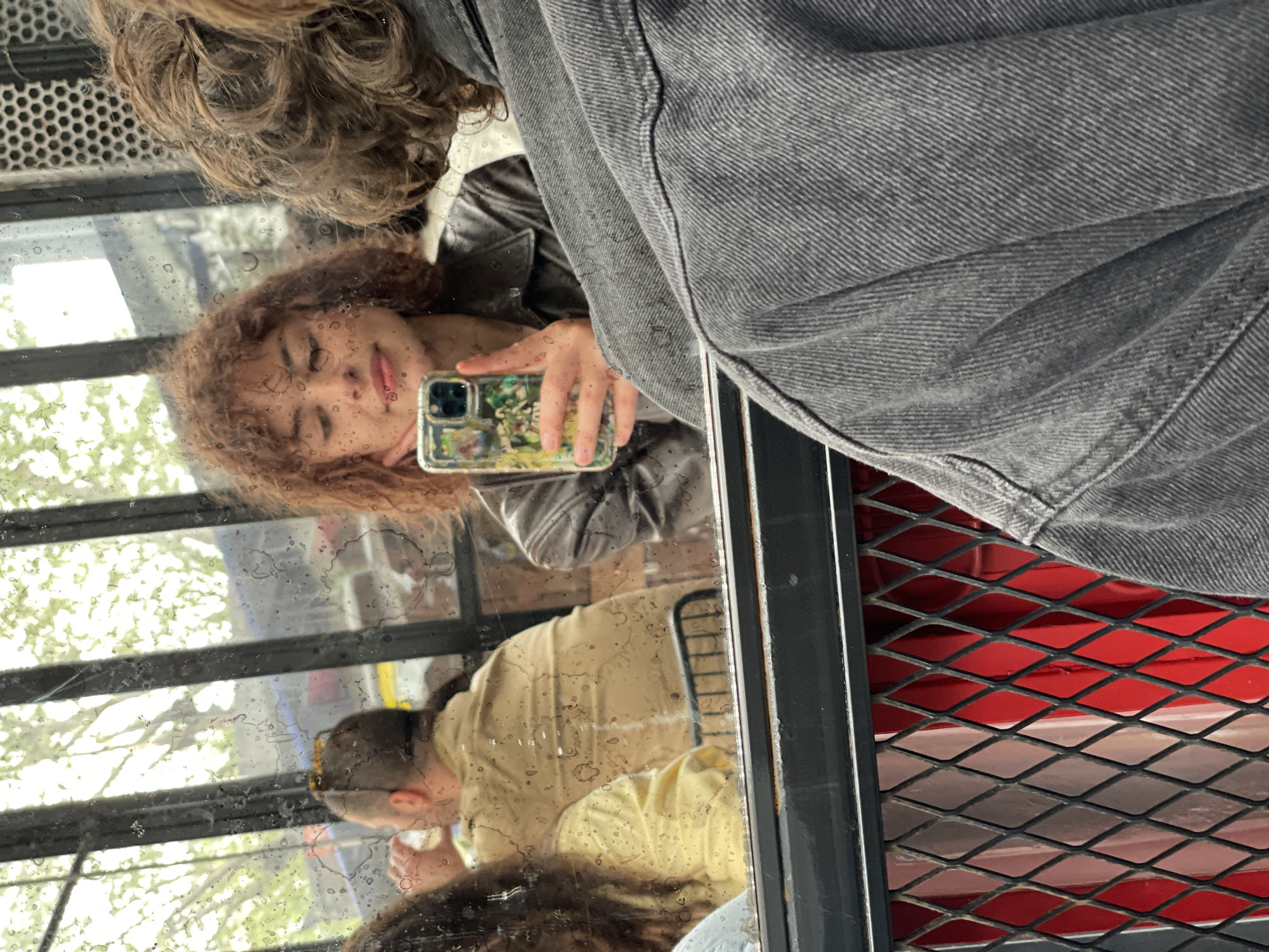 A person is taking a mirror selfie with a cell phone with a green case. The background shows a glimpse of an exterior scene with trees and another person wearing a yellow shirt. A red, metal grid is visible in the lower right corner. The location is not identifiable as Kadıköy, Turkey, based on the visible elements.
