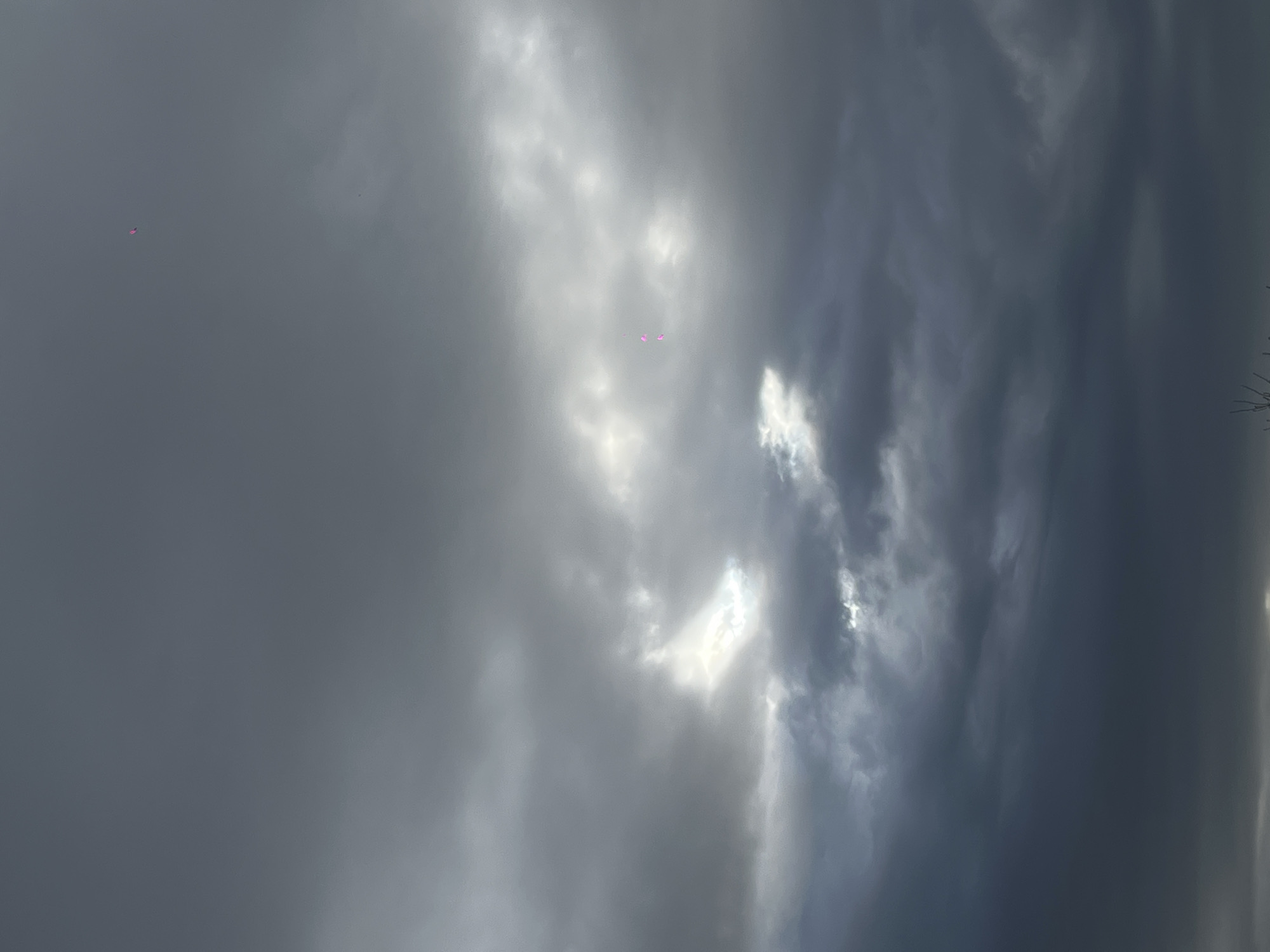 The image displays an overcast sky, rotated approximately 90 degrees clockwise from what appears to be its natural orientation. The sky is filled with a mixture of dense, dark grey clouds and brighter, illuminated cloud formations where sunlight partially breaks through. Patches of intense white light are visible where the sun's rays penetrate the cloud cover.
Several small, pink or magenta-colored objects are discernible against the cloudy background. One distinct, small, multi-faceted pink object is located in the upper-left area of the frame (when viewed in the original orientation), appearing against a darker grey cloud. Closer to the center, two other similar pink shapes, slightly larger and more rounded, are visible amidst the brighter cloud formations. A very small, faint pink dot can also be seen further to the right. These objects resemble balloons drifting in the sky.
The scene captures a daytime setting with significant cloud cover, suggesting either an approaching storm or a generally overcast day. There are no identifiable landmarks or text visible within the frame, but the context indicates the location is Krasnodar, Russia. The presence of multiple pink objects implies an event or celebration involving balloon releases.