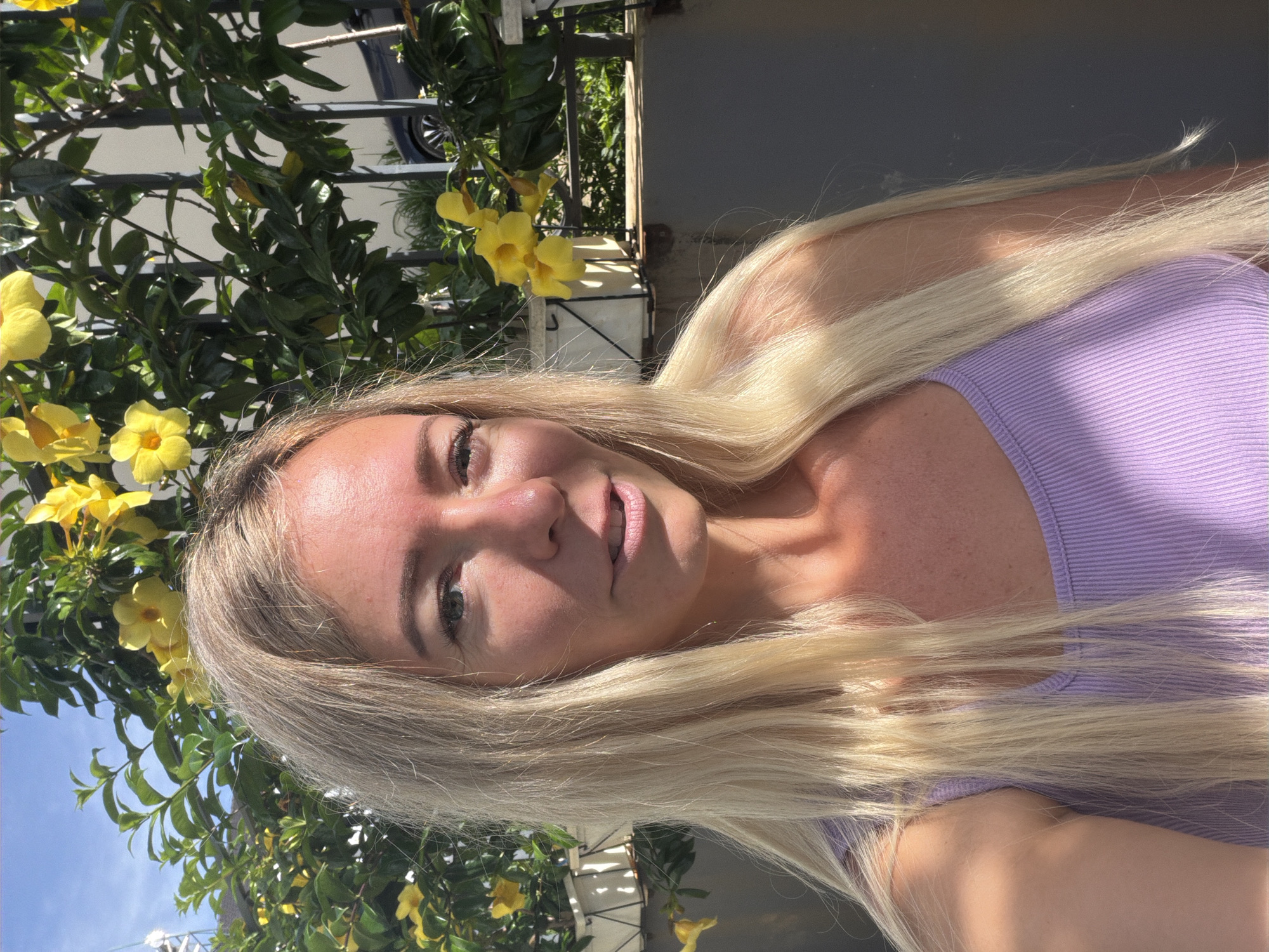 A close-up, head-and-shoulders selfie features a young Caucasian woman with long, straight blonde hair and fair skin. She is wearing a purple ribbed tank top and is positioned outdoors. Behind her, a dense green bush with bright yellow flowers is visible, partially obscured by a dark metal fence. The background also shows a light-colored building and a blue sky. The lighting suggests a sunny day, with sunlight casting shadows on the woman's shoulder and hair. The image is taken in a relaxed, casual setting, possibly a garden or balcony. The woman is looking towards the camera with a slight smile. There are no visible indicators of the location being specifically in Da Lat, Vietnam, or any urban features that would confirm it as such.