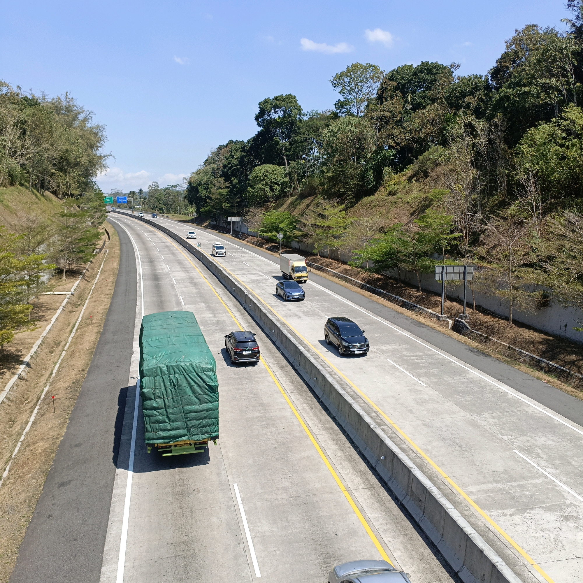 The image shows a highway in Nyamat, Indonesia, on a clear sunny day. The highway has multiple lanes and is flanked by a grassy embankment on the left and a densely forested hillside on the right. Several vehicles are visible on the highway, including a large green-covered truck in the foreground, a yellow truck, a blue sedan, and two black SUVs. There are also a few white cars and vans further in the distance. Traffic signs are visible on the left side of the highway. The overall scene depicts a typical day on a highway in a lush, natural environment.