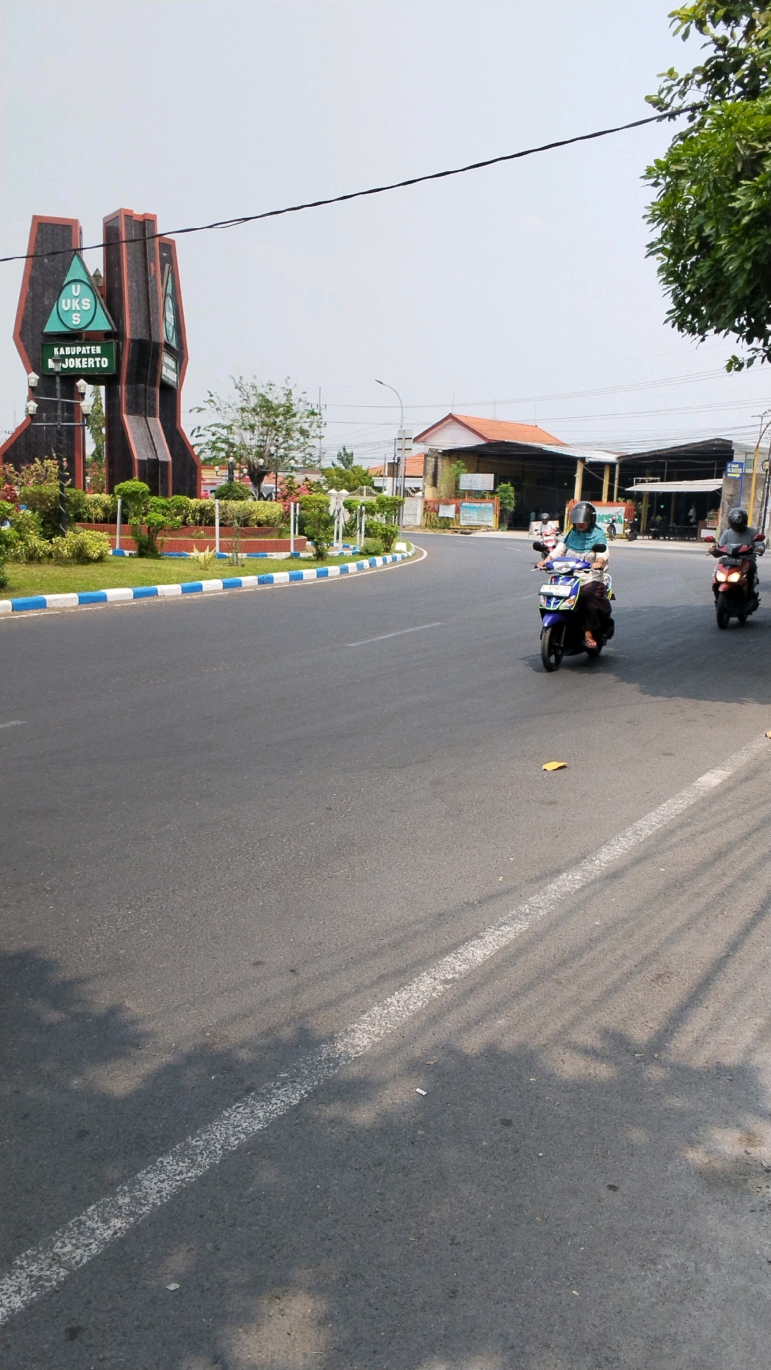 The image shows a roundabout with a monument in the city of Banjaragung, Indonesia. The monument is a large, abstract structure with a green triangular emblem on it. The emblem has a white 'U', 'S', and 'K' arranged in a triangular formation, with the word "KABUPATEN" above "JOKERTO" below. The roundabout is paved with asphalt and has white and blue striped curbing. There are several trees and manicured bushes around the monument. Two individuals are riding motorcycles on the road. The person on the left is riding a blue scooter and is wearing a light blue shirt and dark pants, with a helmet. The person on the right is riding a red motorcycle and is wearing a gray shirt and dark pants, also with a helmet. The sky is clear and bright. Buildings, including a structure with an orange roof and a longer, open-sided building, are visible in the background. The sun is casting shadows of tree branches onto the road.