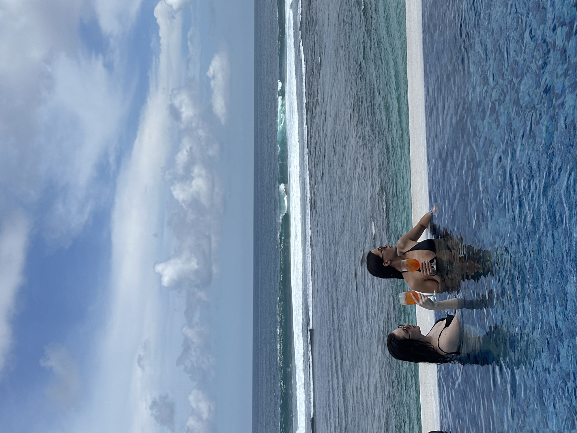 Two women are in an infinity pool overlooking the ocean. The sky is mostly blue with some white clouds. The ocean has white waves. The women are both wearing black bathing suits and sunglasses, and they each hold an orange beverage in a glass. The pool is tiled in a blue mosaic. The scene appears to be in Kuta Selatan, Indonesia, given the typical infinity pools and ocean views in the area.