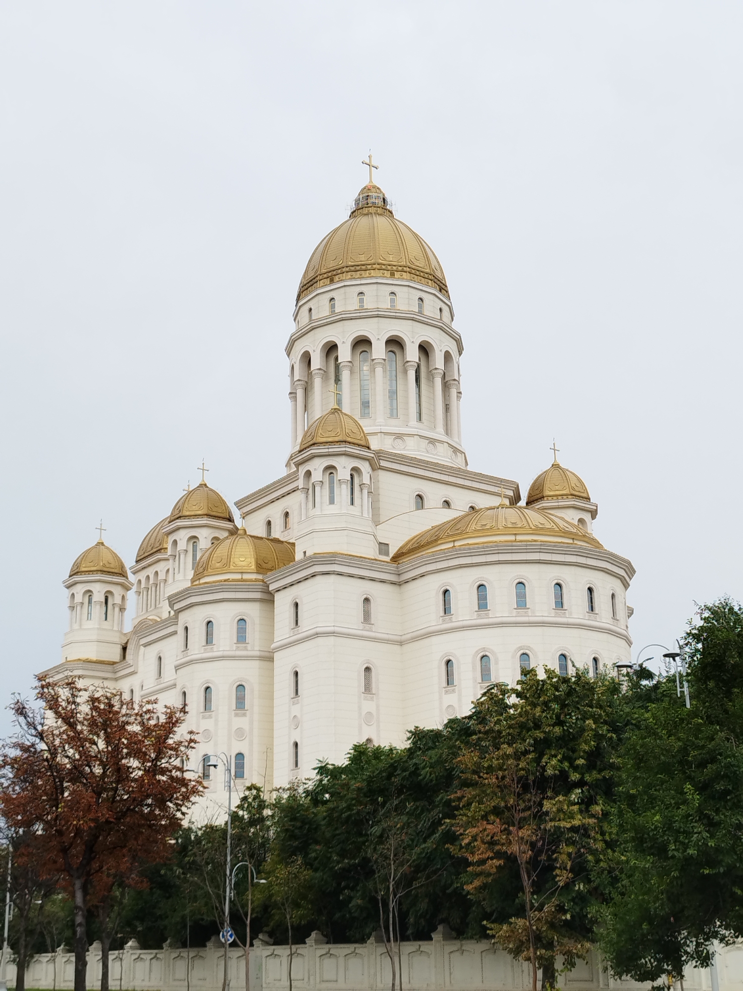 This image shows the People's Salvation Cathedral in Bucharest, Romania. It is a large, white Orthodox Christian cathedral with multiple golden domes, the largest of which is topped with a cross. The cathedral is multi-tiered, with numerous arched windows. In the foreground, there are trees with some autumn foliage, a white decorative fence, and streetlights. The sky is overcast.