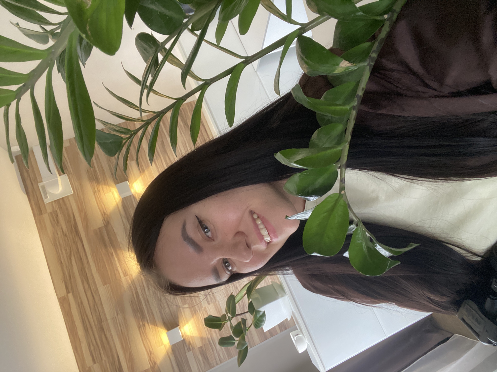 A young woman with long, dark hair and dark eyes smiles at the camera, which is positioned slightly above her. She is wearing a dark brown jacket over a light-colored top. She has winged eyeliner and her teeth are visible in her smile. Behind her, a wooden-paneled wall is illuminated by two modern wall sconces. To her left, a rubber plant in a white pot sits on a white surface, with a small white teacup next to it. To her right, another green plant, possibly a ZZ plant, obscures part of her face and the background. The overall impression is of a casual indoor photograph, likely taken in a residential setting, possibly in Chaikovsky, Russia, given the context.