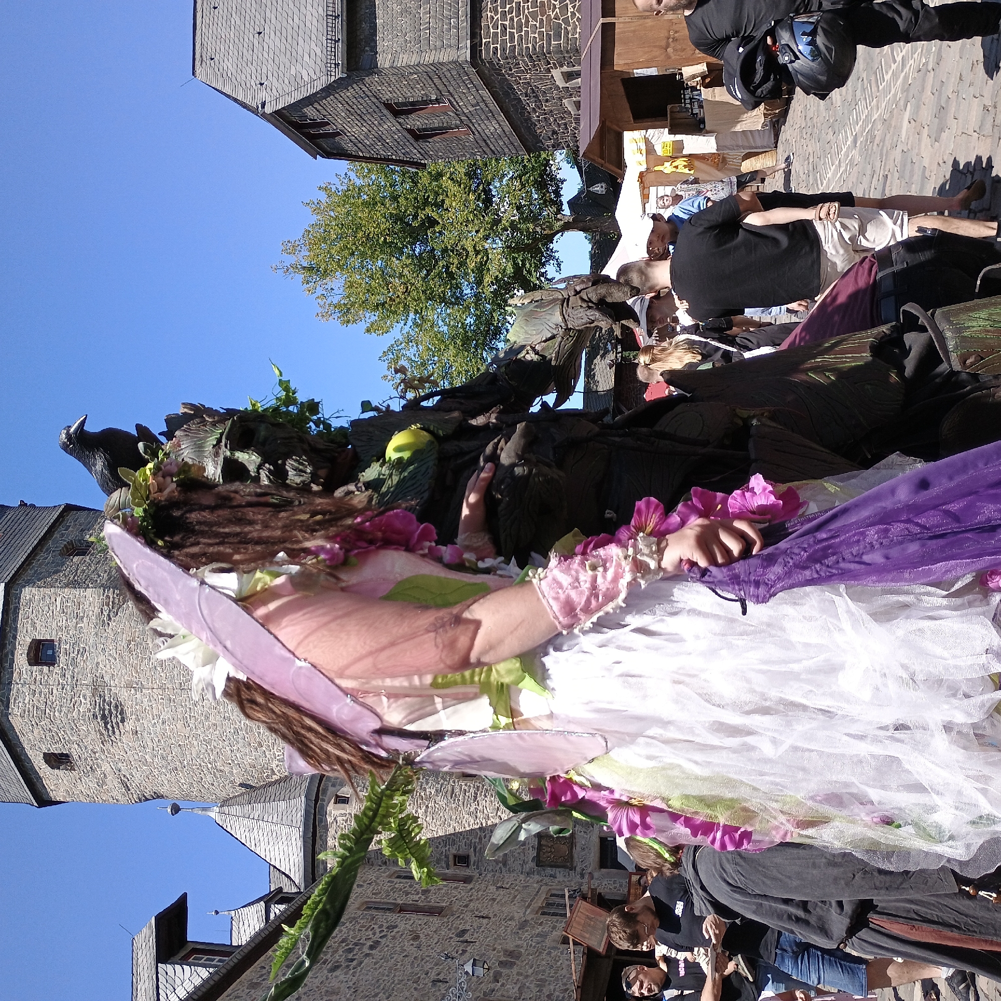The image depicts an outdoor scene in the city of Altena, Germany, likely during a medieval-themed event or festival. In the foreground, a person in a fairy-like costume with a white and pink dress and ethereal wings stands prominently. To the right of the fairy, a large, imposing figure dressed as a "Green Man" or tree spirit is visible, adorned with leaves and branches, and featuring a bird perched on its head. Behind these figures, a cobblestone street is lined with historic stone buildings, including a prominent tower, characteristic of Altena's architecture. Several people are observed in the background, some dressed in casual modern clothing and others possibly in costume, mingling and observing the festivities. The atmosphere appears lively, with market stalls visible in the distance, suggesting a public gathering. The sky is clear and blue, indicating fair weather.