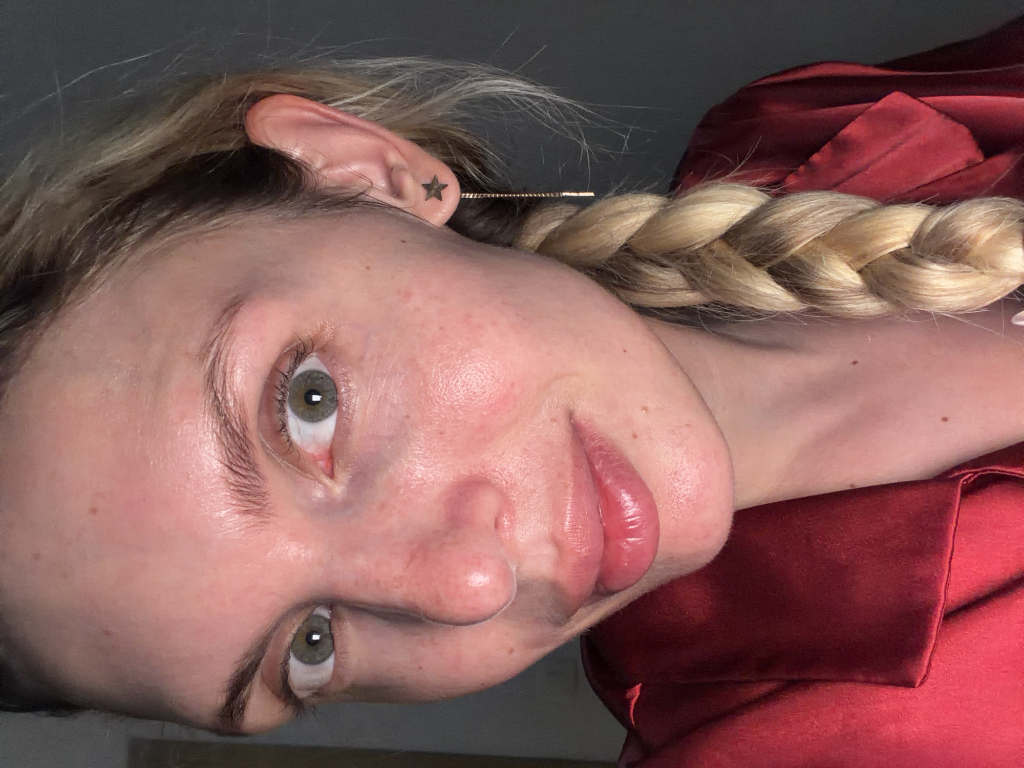 A close-up portrait captures a woman with blonde hair styled in a braid, positioned to the right side of her head. She is seen from the chest up and tilted to the right. A small star tattoo is visible on her left ear. Her skin is fair and appears to be slightly flushed. She is wearing a red silky garment. The background is a neutral gray. The location of this image is unknown and cannot be determined.
