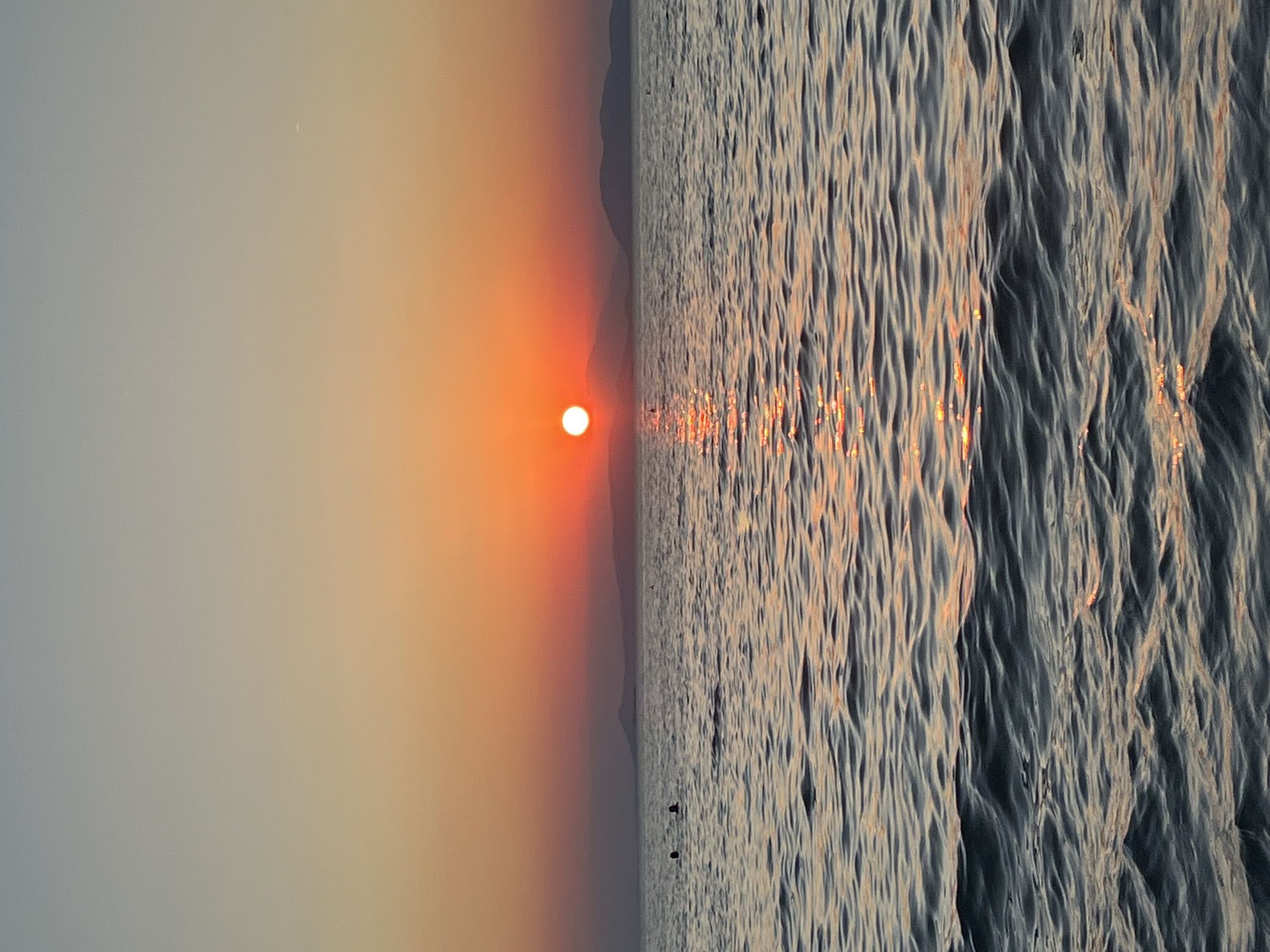 This image depicts a sunset over the sea in Vouliagmeni Municipal Unit, Greece. The sun, a bright orange orb, is low on the horizon, casting a shimmering reflection on the rippling water. The sky transitions from a pale gray at the top to a warm orange and red near the horizon. In the distance, a silhouetted mountain range is visible. Two figures can be seen swimming in the water, their heads just above the surface. The foreground is dominated by the textured surface of the sea, with gentle waves catching the light.