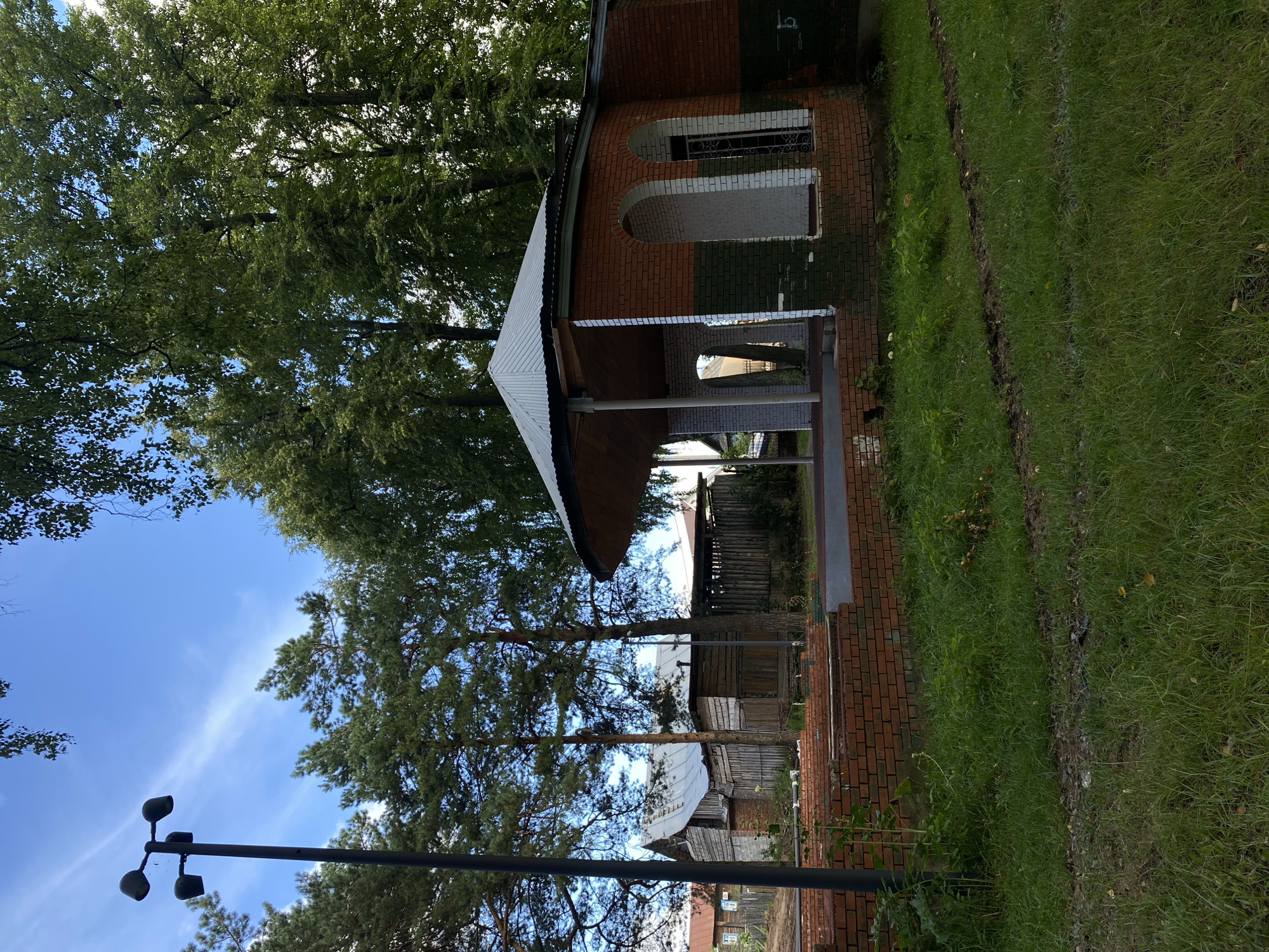 The image shows a view of the Votkinsk City Park, specifically the area around the open-air stage. A partially constructed brick structure with arched openings and a metal roof stands in the foreground. In the background, there are several wooden sheds and buildings, characteristic of rural Russian architecture. Tall trees, including pines and deciduous trees, form a lush green backdrop against a blue sky with scattered clouds. A lamppost is visible to the left of the frame. The ground is covered in grass, with some areas of bare earth and tire tracks. The overall scene depicts a tranquil park setting with elements of both natural beauty and rustic man-made structures.