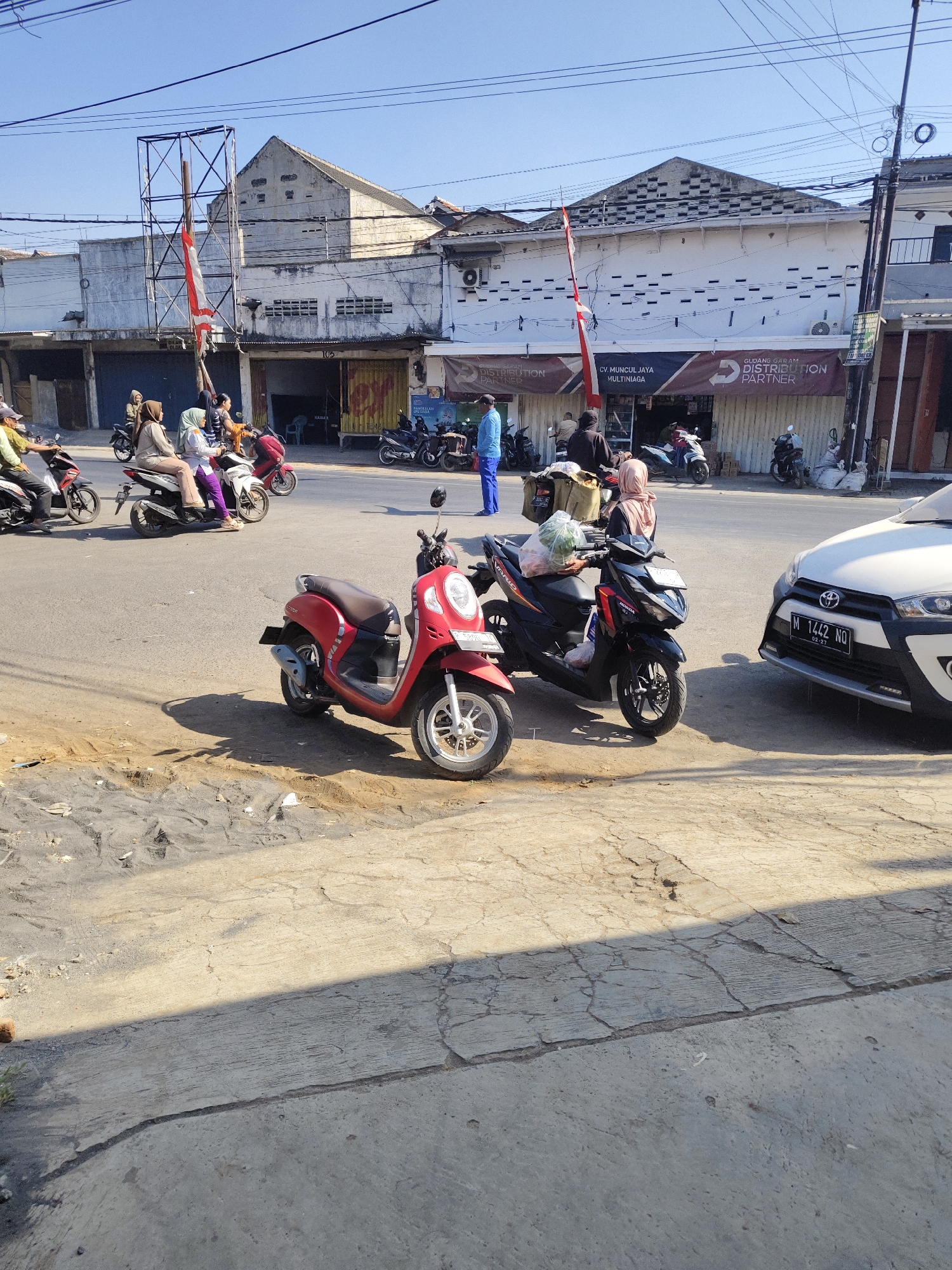 The image captures a street scene in Gambirono, Indonesia, under a clear blue sky. In the foreground, two scooters are parked on a dusty, cracked concrete surface, with a shadow cast across it. A red scooter is parked to the left, and a black scooter with items strapped to the back is to the right. Beyond the scooters, a white car is parked. The street is paved and shows signs of wear.
In the middle ground, several people are visible on motorcycles and standing near a building. A man in a blue shirt and pants stands near a black motorcycle. A woman wearing a hijab and a light-colored jacket is seated on a white scooter, while another woman in a pink hijab is standing next to her. Other individuals on motorcycles are also present.
The background features a row of buildings, some of which appear to be shops or businesses. One building has a white facade with dark horizontal windows and a banner that reads "GUDANG GARAM DISTRIBUTION PARTNER" and "CV. MUNJUL JAYA MULTINIAGA". Another building to the left has a more weathered appearance with a large, empty metal frame structure in front of it, on which a Indonesian flag is hanging. Overhead, power lines crisscross the sky. The overall impression is of a busy, everyday street scene in an Indonesian town.