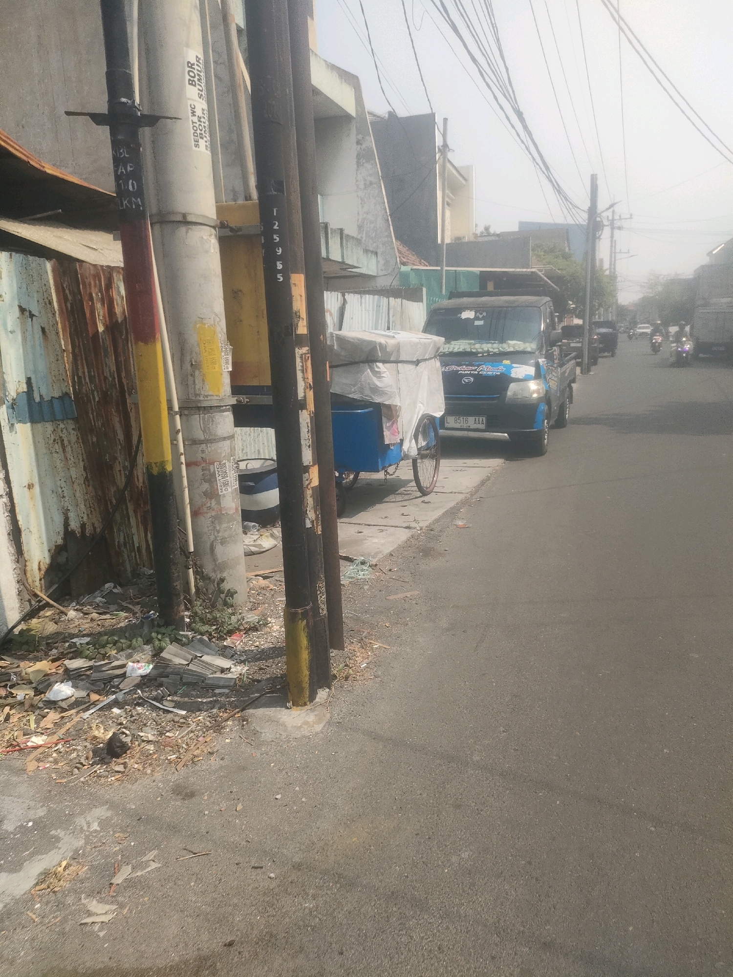 This is a street scene in Surabaya, Indonesia, on a hazy day. A blue and black pickup truck, with license plate L 8516 AAA, is parked on the right side of the road. In front of the truck, a blue cart covered in white fabric is also parked. The street is paved with asphalt and there is debris and some vegetation along the roadside. Several utility poles stand along the sidewalk, some of them marked with numbers or text. In the background, there are buildings and more utility poles with a tangle of wires. A few motorcycles are visible further down the street.