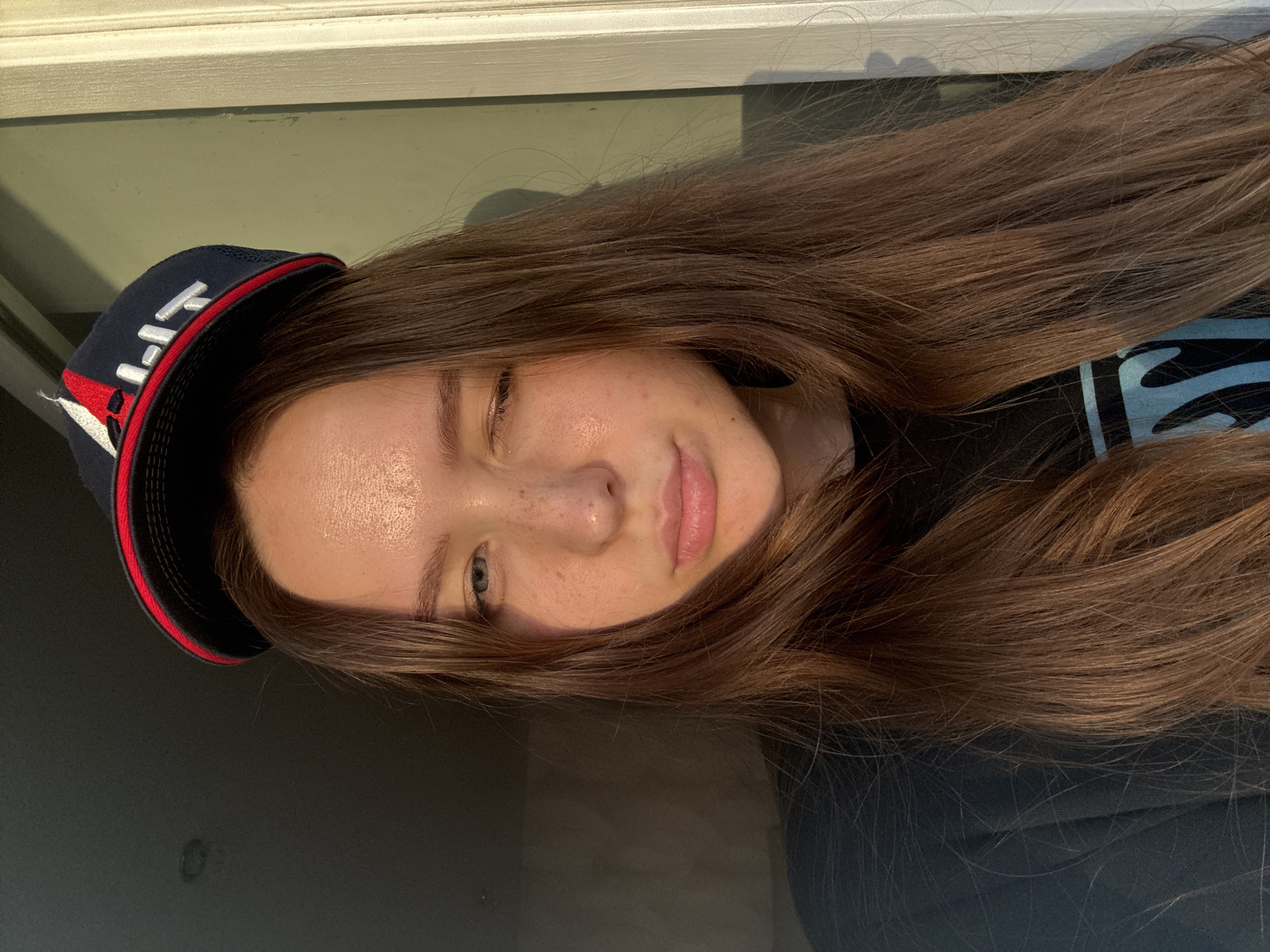 A rotated, close-up portrait shows a young woman indoors in Chaikovsky, Russia. She is looking towards the camera with a playful expression, one eye slightly squinted or winking, and her lips gently parted. Her long, straight, brown hair flows across the frame.
She wears a dark blue baseball cap with white embroidered text, which appears to be "HPT," and a red stripe along the brim. Her skin is fair, with visible freckles on her nose and cheeks. Her eyes appear blue or greenish. Strong, natural light illuminates her face from the left, suggesting it is daytime.
She is wearing a dark-colored top with a light blue or white abstract pattern visible on her right side. In the background, above her head, a white architectural frame, possibly a window or door frame, is visible against a muted green or gray wall. The lower portion of the background is dark, likely a shadow or a dark surface.