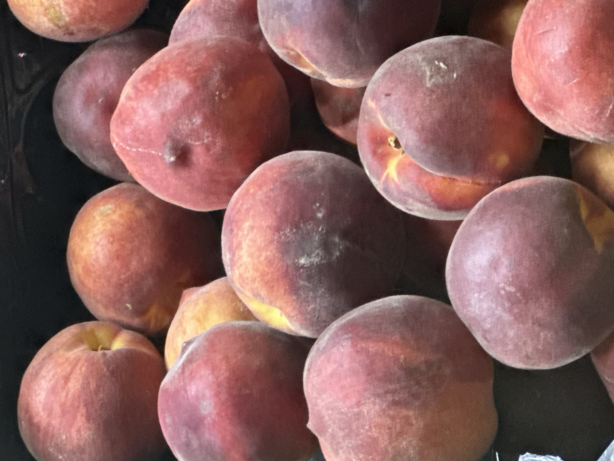 A close-up, overhead shot shows a dark, cluttered display of several ripe peaches. The peaches are round and fuzzy, with skin tones ranging from deep red to yellow and orange. They are packed together, with some peaches overlapping others. The background is dark and out of focus. The image appears to be taken in an outdoor market or grocery store. The specific location in Sochi, Russia cannot be determined from the image alone.