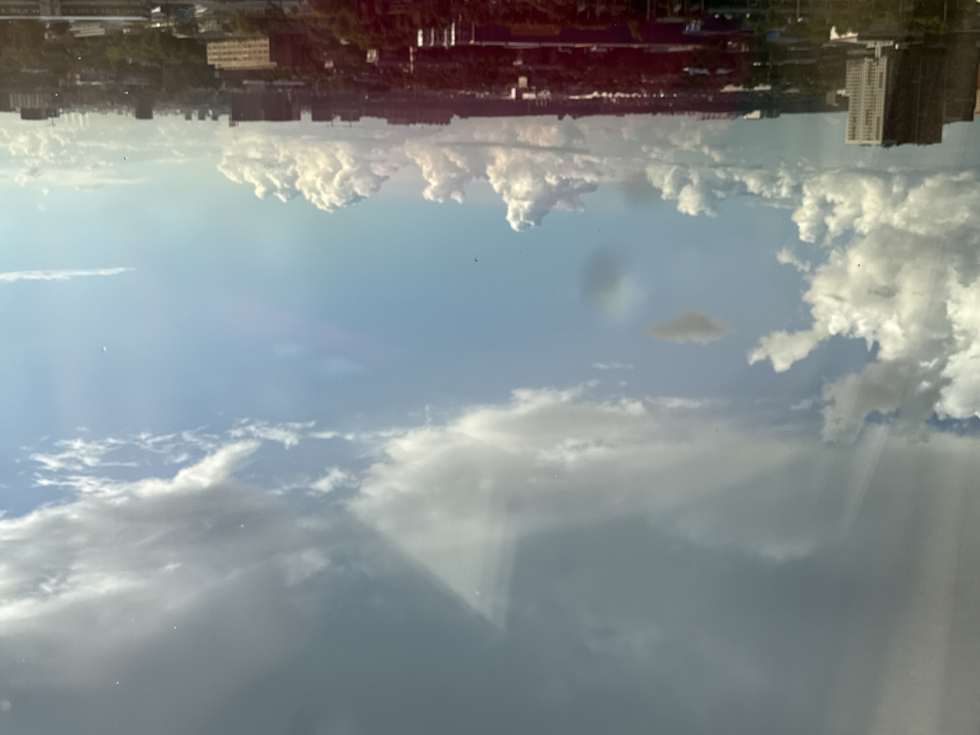 This image captures a wide-angle view of a city skyline from an elevated perspective, likely a window. The sky dominates the upper portion of the frame, filled with a mix of dramatic, heavy gray clouds and lighter, wispy white clouds, suggesting varied weather conditions. Sunbeams appear to be breaking through some of the darker clouds, casting rays across the sky.
The lower half of the image displays a dense urban landscape, characterized by numerous buildings of varying heights and architectural styles. Tall, modern apartment blocks with light-colored facades are visible on the left side. In the distance, a sprawling network of lower-rise buildings, interspersed with greenery, extends towards the horizon. Several construction cranes are also visible, indicating ongoing development within the city. The overall impression is of a busy, developed metropolitan area under a dynamic sky. The scene is likely in Lyubertsy, Russia, given the context. There are no visible people or direct interactions in the frame. The environment is urban, with a significant focus on the sky and city architecture.