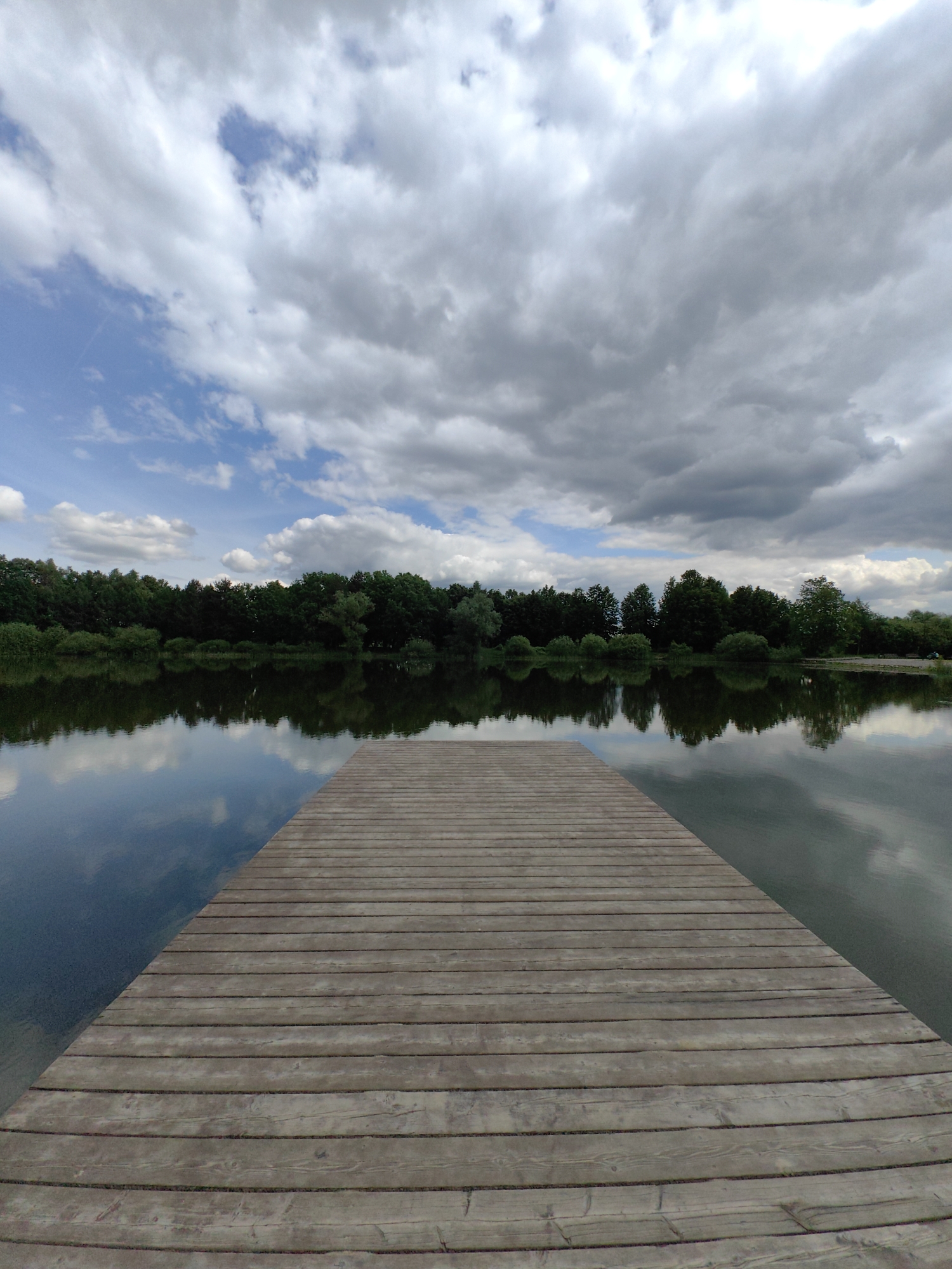 A medium-angle, eye-level wide shot shows a wooden dock extending into a calm lake, reflecting trees and a partly cloudy sky. The dock is made of horizontal wooden planks running the length of the dock toward the center. The lake's surface is relatively smooth, with reflections of the trees and sky creating an almost mirrored effect. A line of trees forms the far shore of the lake, consisting of a mix of leafy green trees. The sky is filled with white and grey clouds, with patches of blue sky visible between them. The clouds are reflected in the lake's surface. The image suggests a tranquil natural scene. The location is Domodedovo, Russia.
