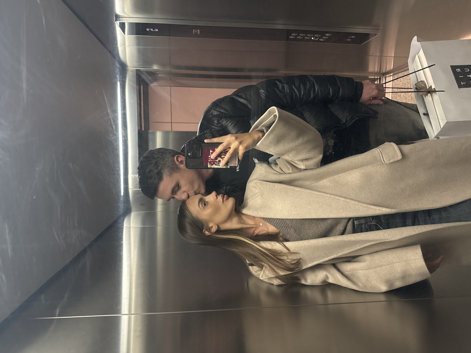 A couple is captured in a selfie taken inside a mirrored elevator. The man, wearing a black puffer jacket, kisses the woman's forehead. She is dressed in a gray sweater and a light beige coat with prominent lapels. A shopping bag with dark sticks is visible to the side. The reflections create a dynamic perspective, showcasing the modern interior of the elevator with its stainless steel surfaces and controls.