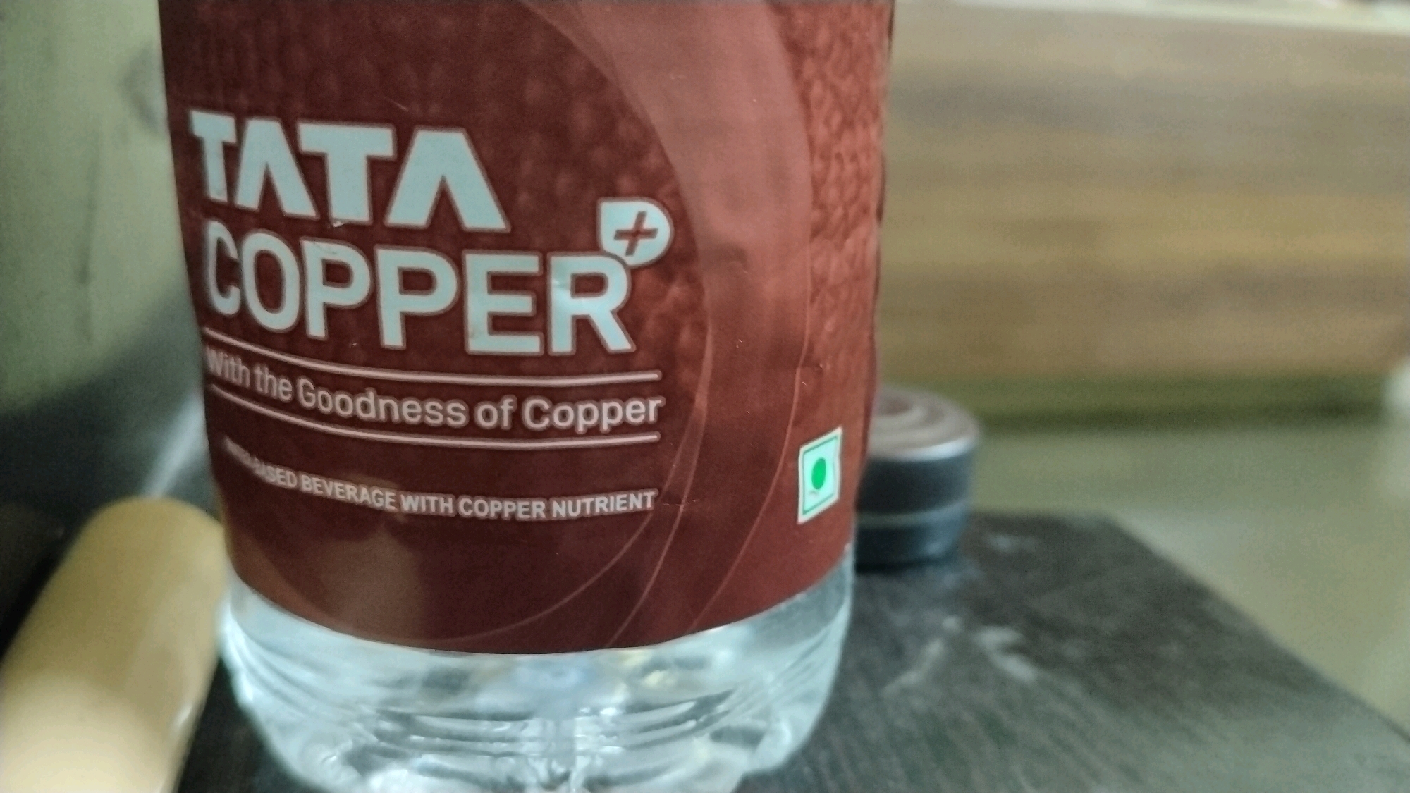 The image shows a close-up of a bottle of "TATA COPPER" water. The label is primarily brown with white text. The text "TATA COPPER" is prominently displayed in large, white, sans-serif font. Below this, in a smaller font, it reads "With the Goodness of Copper". Further down, the text "SED BEVERAGE WITH COPPER NUTRIENT" is partially visible. A small green circle inside a square, indicating a vegetarian product, is also visible on the label. The bottle itself appears to be clear plastic with visible water inside.
In the background, a blurred wooden surface can be seen, possibly a table or shelf. To the left of the bottle, a light-colored, cylindrical object, possibly a pen or a similar item, is partially visible. To the right of the bottle, a dark-colored, circular object with a ridged edge, possibly a cap or a small container, is also blurred. The overall setting suggests an indoor environment, possibly a desk or a countertop.
There is no specific information in the image that would indicate its location in Hyderabad, India. The image focuses solely on the product and its immediate surroundings.