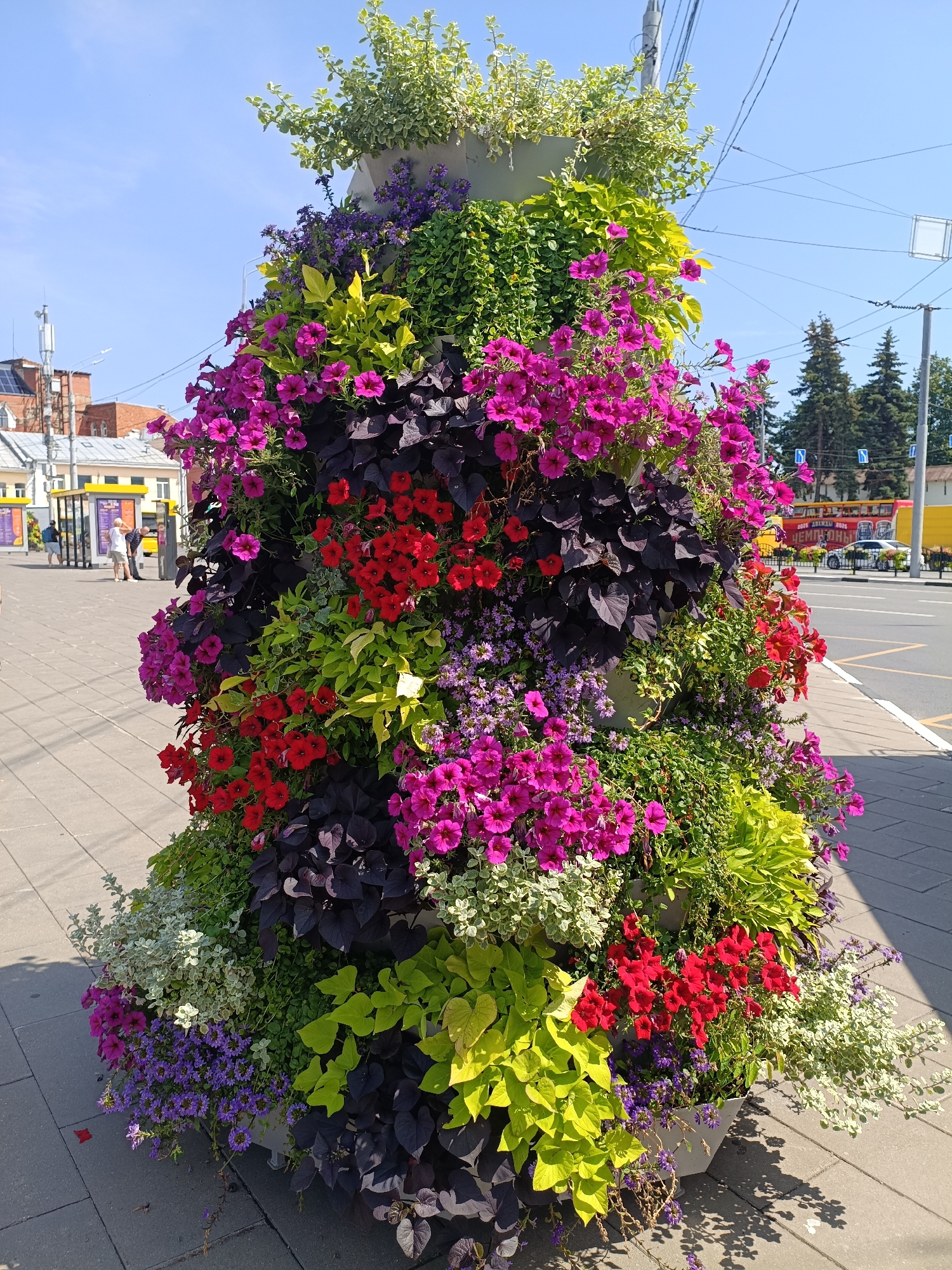 A tall, tiered planter overflowing with colorful flowers and foliage stands on a paved sidewalk in the city of Yaroslavl, Russia. The planter is densely packed with a variety of plants, including bright pink petunias, red impatiens, deep purple iresine leaves, chartreuse sweet potato vines, and small purple flowers, all cascading down its sides. In the background, a street runs alongside the sidewalk, with buildings, traffic signals, power lines, and trees visible. A yellow bus stop shelter with people waiting and a passing car are also present. The sky is clear and blue, indicating a sunny day.