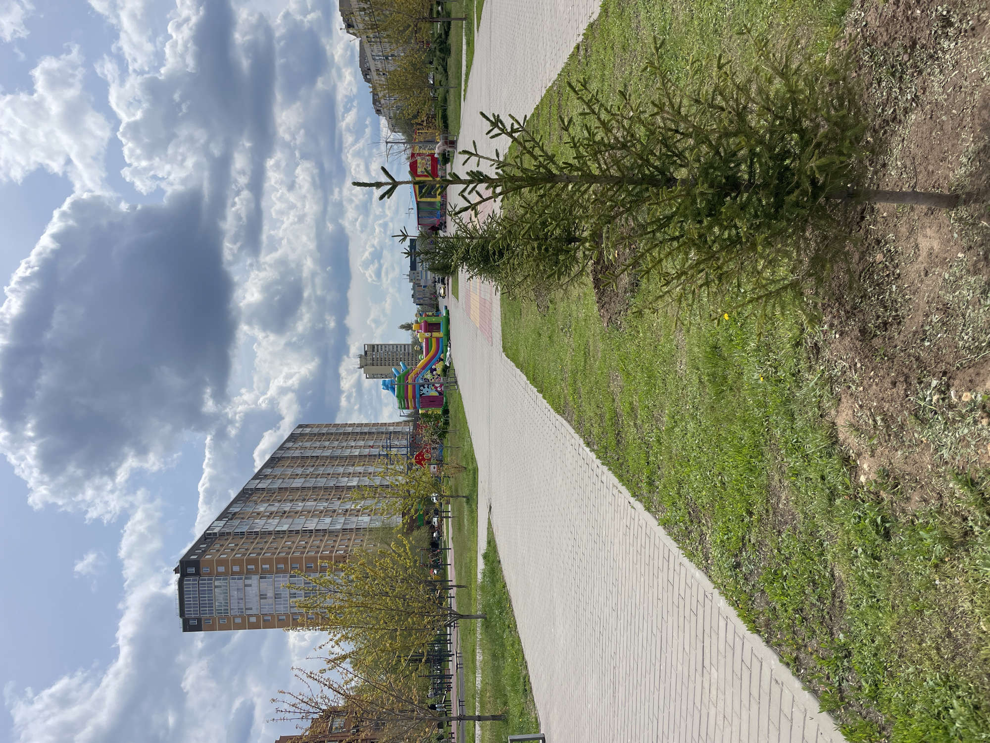 This media file, presented in multiple crops, depicts a paved pedestrian path running through a green area, flanked by grass and young trees. The path is made of light grey interlocking bricks and extends into the distance. On either side of the path, there is green grass, with some areas showing patches of bare soil, suggesting recent planting or landscaping. Several small, green evergreen trees (possibly spruce or fir) are planted along the path, particularly on the right side. Other deciduous trees, some with new spring leaves, line parts of the path and are visible in the background.
In the mid-ground, to the right of the path, a colorful inflatable bouncy castle or playground structure is visible. Another, similar structure can be seen further in the background, near a tall building. This suggests a children's play area or a temporary event.
In the background, on the left side, a modern, tall residential building with many windows dominates the skyline. Another, smaller building is visible further behind it. The sky above is partly cloudy, with large, white cumulus clouds and some darker grey clouds suggesting a dynamic weather pattern, but no immediate threat of rain.
The scene appears to be set in an urban or suburban residential park or courtyard. It is daytime, likely late morning or afternoon, given the bright, natural light. No people are visible, and no specific activity is clearly taking place, though the inflatable structures imply a potential for play. No visible text is discernible in the images. The overall impression is one of a calm, well-maintained public space.