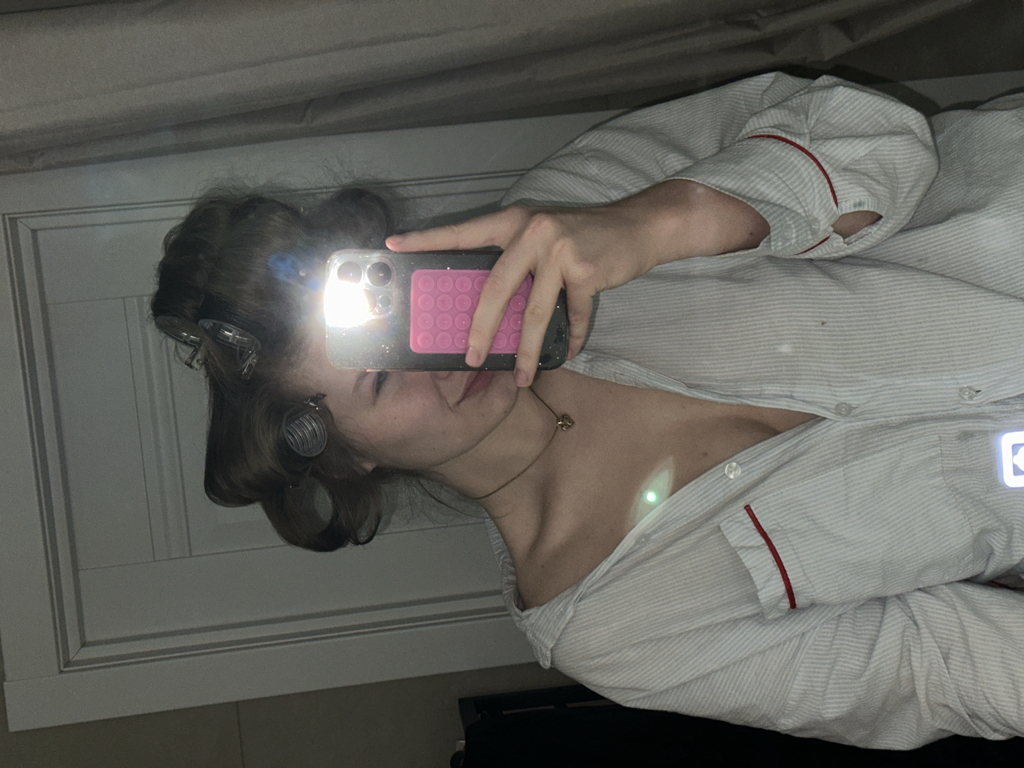 A person is taking a selfie in a mirror. The person is wearing striped pajamas with red trim. Their hair is in curlers. They are holding a smartphone with a pink pop socket on the back. The location is unknown and cannot be determined from the image.