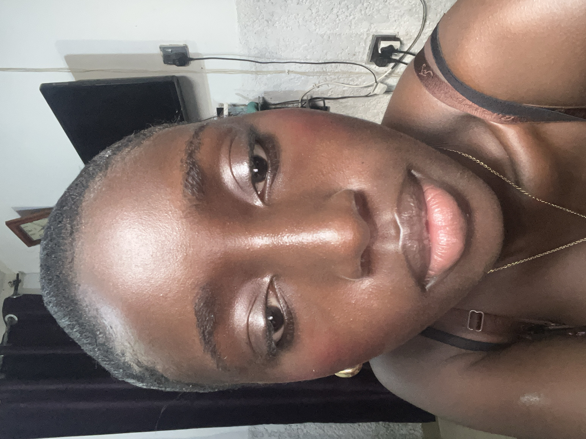 A close-up, portrait-style photograph shows a dark-skinned person with their face positioned prominently in the frame. The person is wearing a thin gold chain necklace and a bra with brown and black straps. A small gold hoop earring is visible on the left side of the frame. The background is somewhat blurred, showing a white wall with electrical outlets and wiring. A television screen and dark curtains are also visible in the background. There is no information to confirm that the photograph was taken in Abuja, Nigeria.
