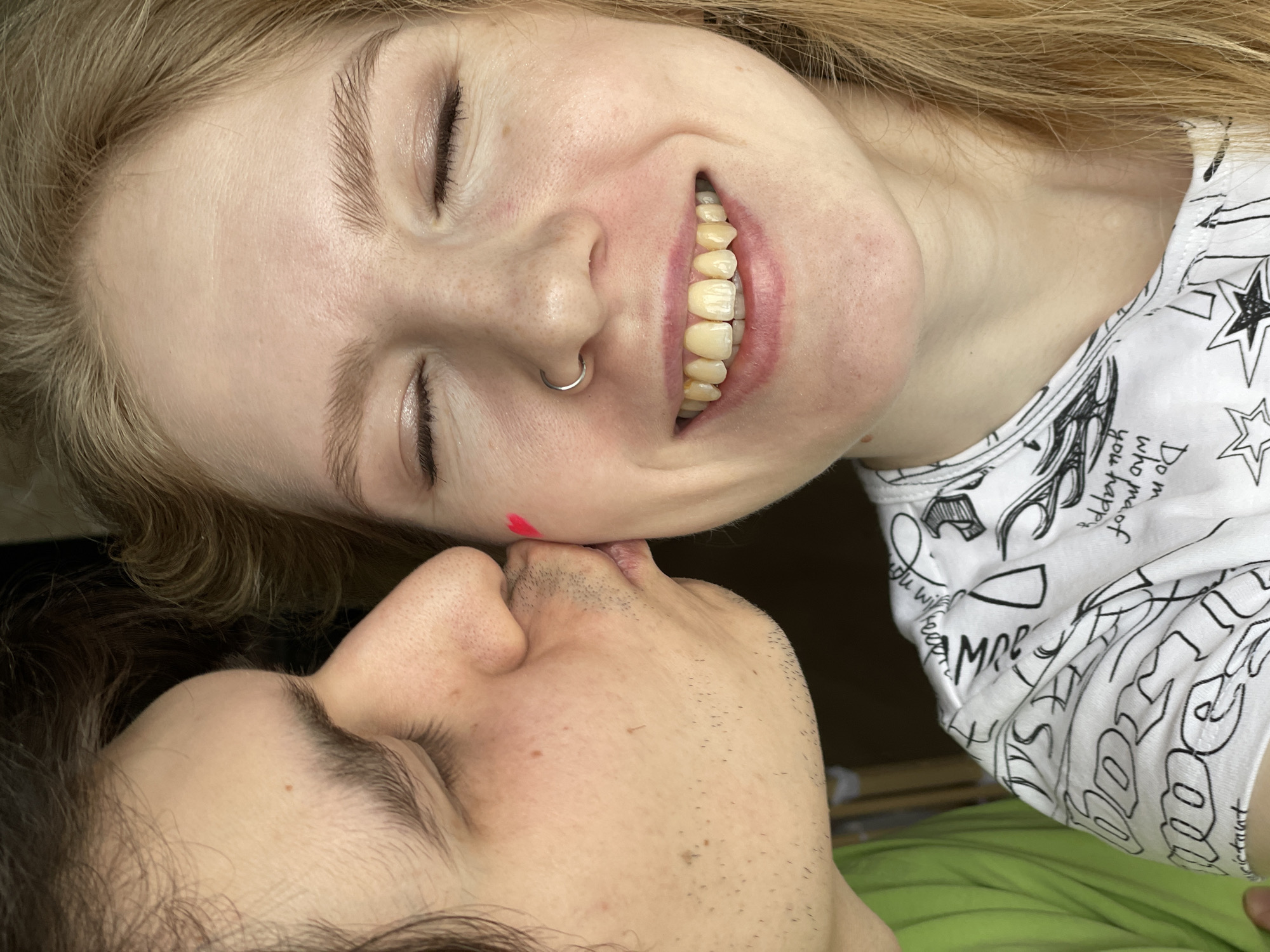 This intimate, diagonally oriented close-up captures two individuals in a moment of affection, appearing to be lying down. The upper subject is a person with long blonde hair, fair skin, and closed eyes, displaying a wide, joyful smile that reveals yellowish teeth. They wear a silver hoop nose ring and have a small, bright red mark, possibly a heart shape or lipstick imprint, on their cheek. Their white t-shirt features black handwritten text and doodles, including partially legible words like "Happy," "love," "AMOR," and "life," along with star graphics.
Beneath them, partially obscured, is a person with dark hair, visible stubble on their face, and closed eyes. Their lips appear to be in gentle contact with the upper subject's mouth or nose area, suggesting a tender kiss or affectionate interaction. The background is dark, with a hint of bright green fabric, possibly a blanket, at the bottom. Both individuals' closed eyes and the prominent smile convey strong emotions of happiness and closeness.