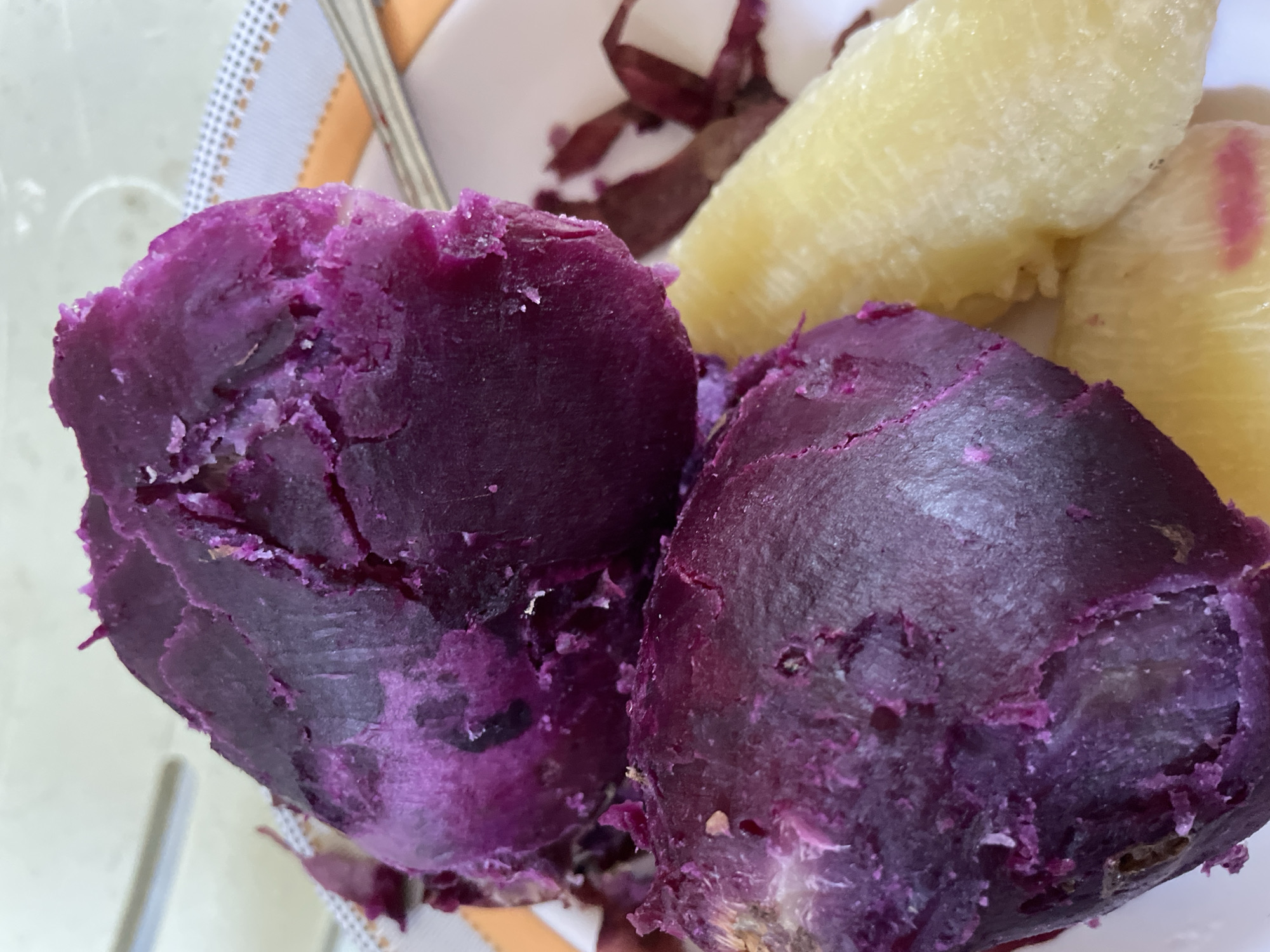 The image displays a close-up view of two purple sweet potatoes, likely Ube, on a white plate with an orange and white checkered border. The sweet potatoes appear to have been peeled and possibly steamed or boiled, as their texture is soft and slightly cracked, revealing a vibrant purple interior. To the right of the purple sweet potatoes, there are pieces of what appears to be plain cooked potato or a similar starchy vegetable, with a pale yellow color. A silver fork is partially visible on the right side of the plate. The background is blurred, suggesting an indoor setting, possibly a dining area. There is no discernible information within the image to indicate its specific location in Davao City, Philippines.