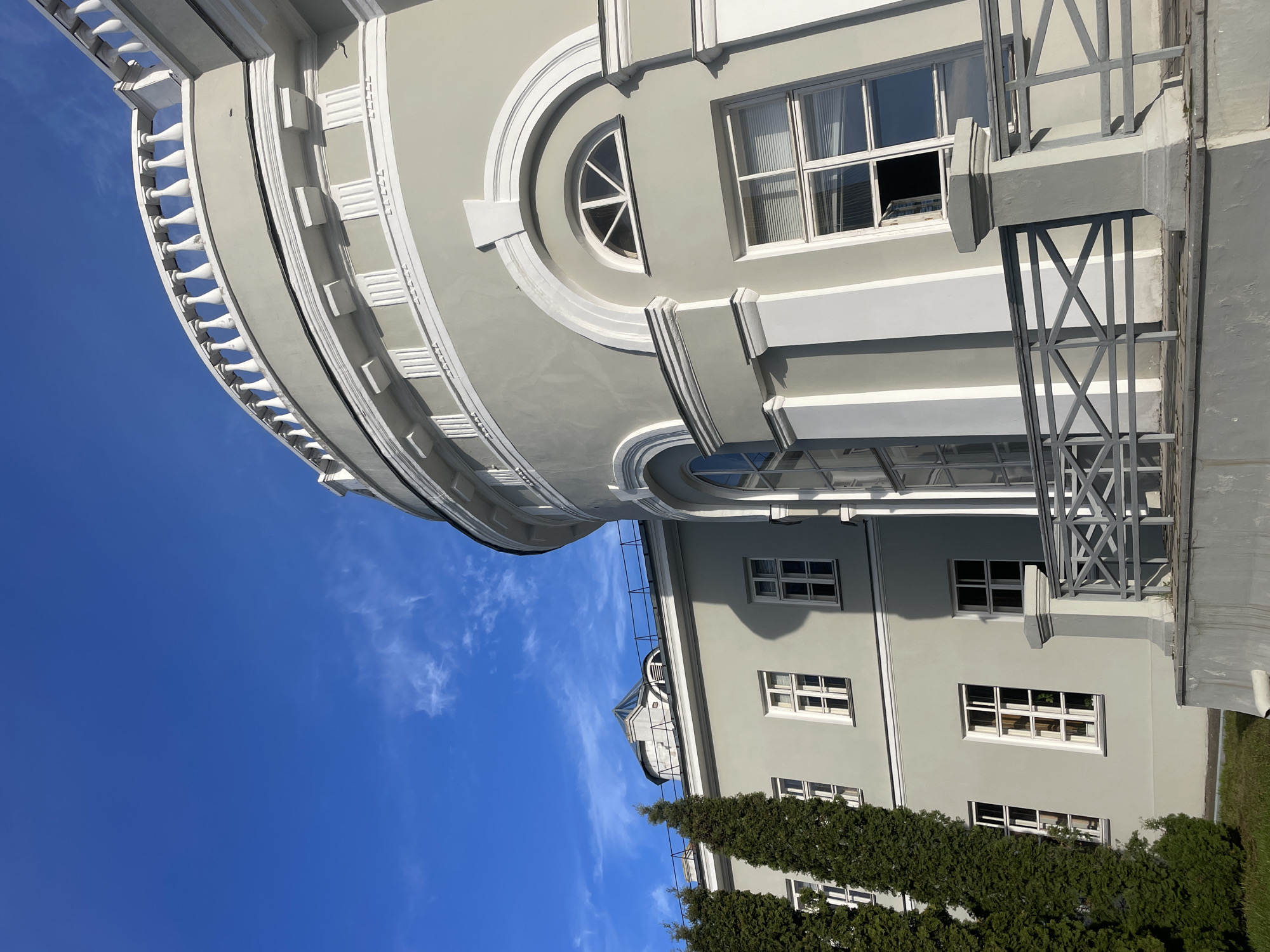 The image displays a multi-story building, likely located in Kazan, Russia, characterized by its classical or neoclassical architectural style. The main subject is a light grey facade adorned with crisp white decorative elements, set against a clear, vibrant blue sky with a few wispy white clouds.
The building features a prominent curved section at the top, bordered by a white balustrade with evenly spaced balusters. Below this, the facade includes decorative rectangular blocks along a white molded edge. A notable architectural detail is a semicircular window (lunette) framed by an ornate circular molding, positioned between larger rectangular windows. One of the rectangular windows is slightly open, revealing internal blinds.
To the right, a sturdy grey metal structure, possibly a fire escape, balustrade, or support for an overhang, is visible with a pattern of vertical bars and 'X'-shaped cross-bracing. Lower sections of the building also feature rectangular windows and a visible flat roofline. Further down, dark green coniferous trees or bushes line the base of the structure.
The photograph was taken during daytime under bright sunlight, creating distinct shadows on the building's exterior and highlighting its architectural details. No people are visible, and no specific event or activity is taking place. There is no visible text in the image.