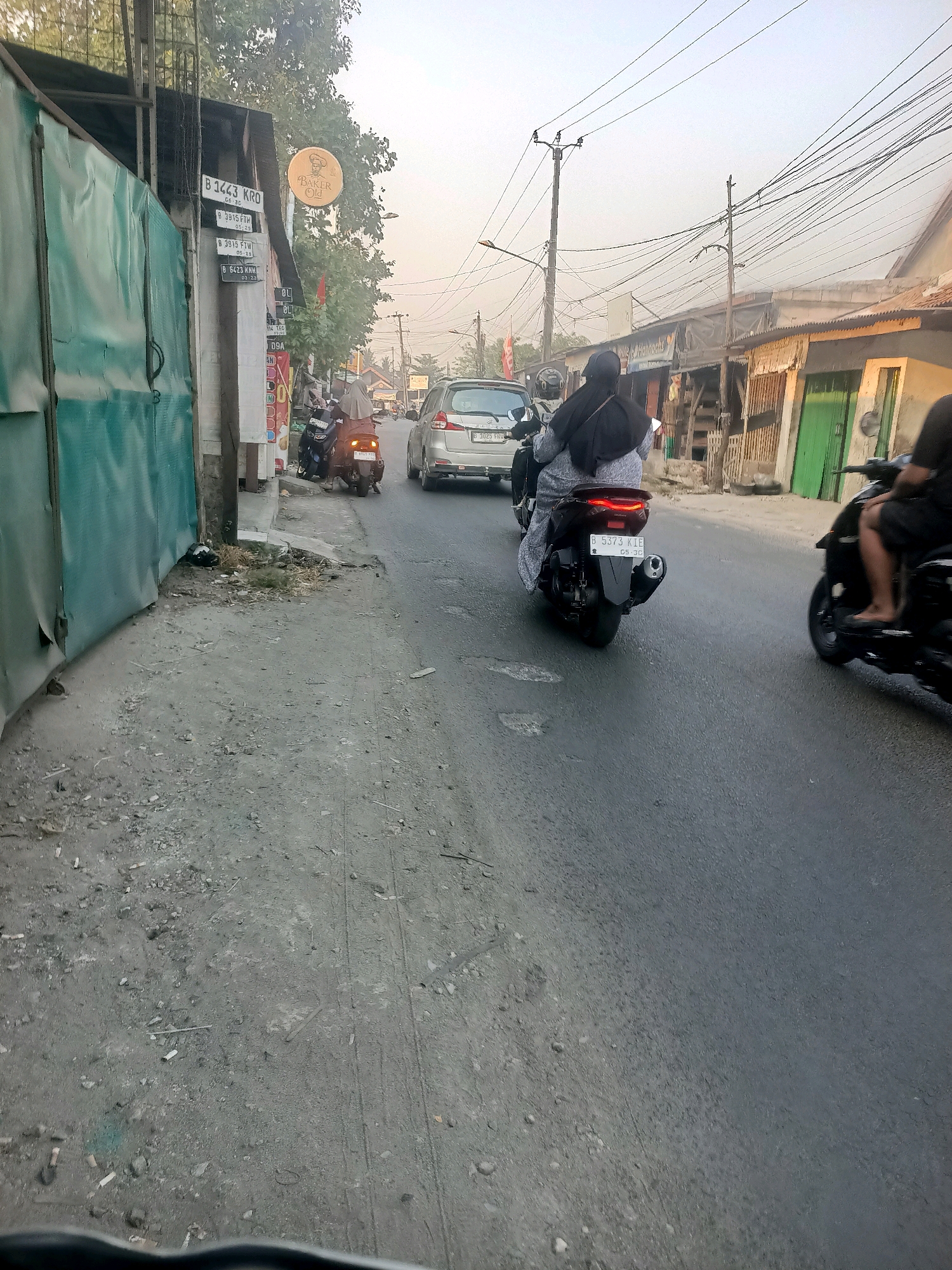 In Wanajaya, Indonesia, a street scene unfolds with a focus on a two-wheeled vehicle and its occupants. The image captures a street with a sidewalk and several buildings along the side. A person wearing a headscarf and a long-sleeved shirt rides a motorcycle, with another individual also wearing a headscarf on the back. They are moving along the asphalt road. In the background, a silver car is visible, and further down the street, another motorcycle with a rider is present. The buildings appear to be shops or residences, with some displaying signs. The sky is hazy, suggesting potential air pollution or dust. The ground on the sidewalk and road shows signs of wear and tear, with scattered debris. The overall impression is a typical urban street environment in Indonesia.