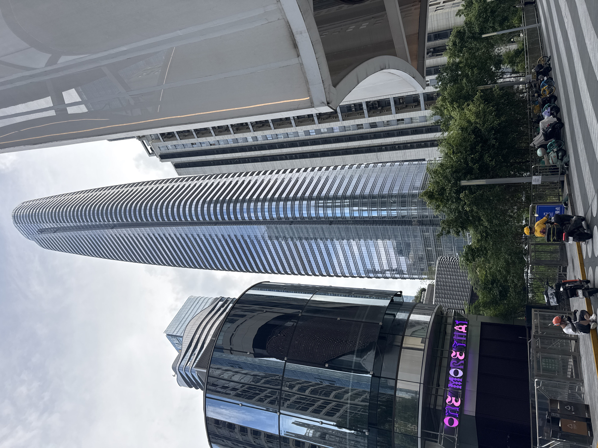 The image captures an urban scene in the Futian District of China, showcasing modern architecture. The focal point is a cluster of tall buildings with reflective glass facades. One skyscraper is distinguished by its elongated shape and evenly spaced horizontal lines. Another building features a curved design. The 'ONE AVENUE' signage is prominently displayed. Trees and sidewalks line the base of the buildings, offering a hint of green amidst the concrete. Two people are visible; one is wearing a yellow shirt and standing near a scooter, and the other is wearing a white shirt, sitting and smoking a cigarette. Various parked vehicles are also visible.