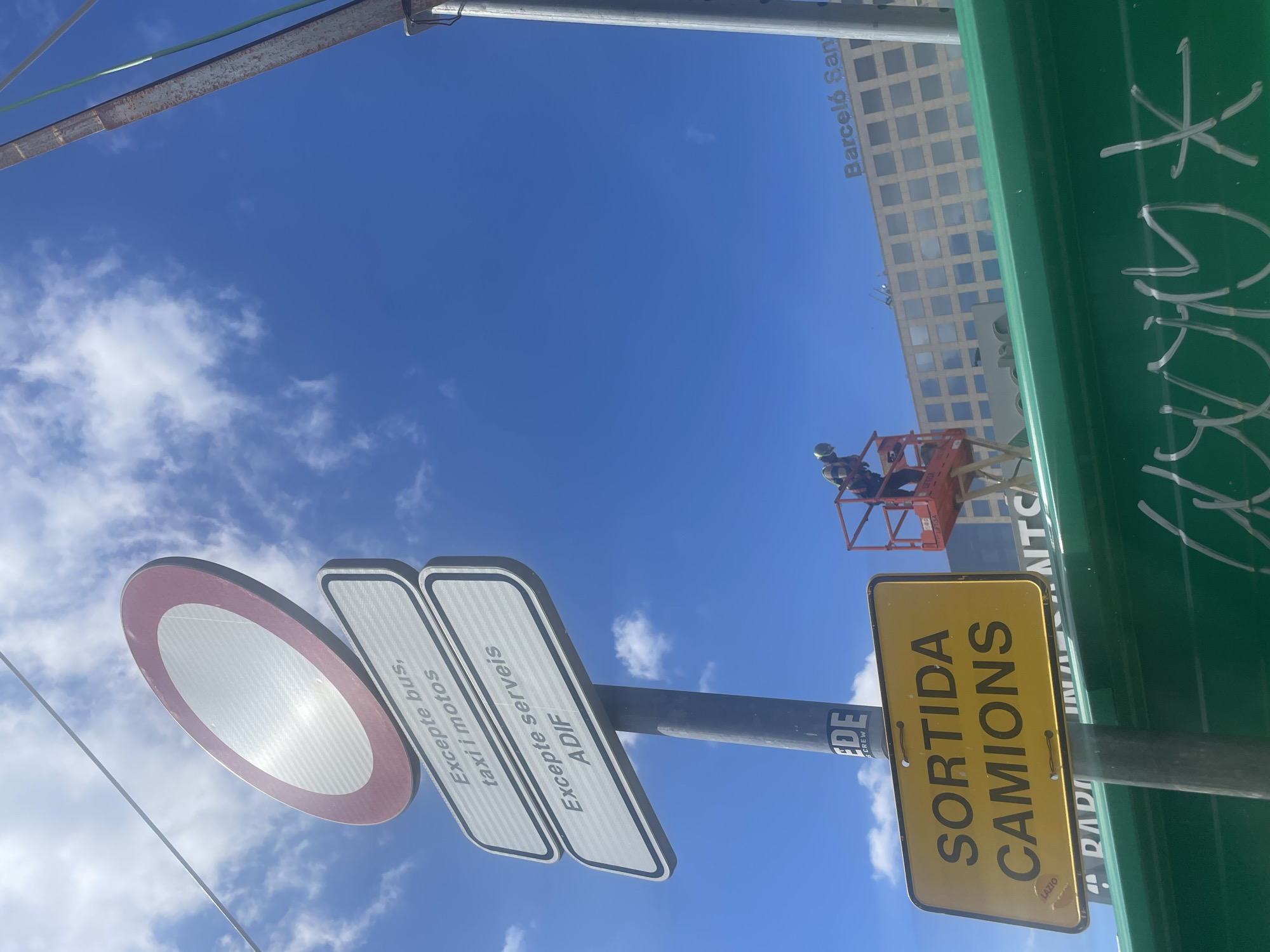 Here is a description of the image.
The image shows a clear blue sky with scattered clouds. The view is from below, looking up at a series of road signs mounted on a gray pole. The signs include a white circle with a red border (no entry), rectangular signs with white backgrounds and gray borders, and a yellow rectangular sign with black text reading "SORTIDA CAMIONS" (Truck Exit) with "LAZIO" at the bottom corner. The other signs have text in Catalan, reading "Excepte bus, taxi i motos" and "Excepte serveis ADIF." The letters "EDE" and "CREW" are also visible on the gray pole.
In the background, a building with a grid-like facade and the text "Barceló San" can be seen. To the right of the building, a person in a safety harness is on a platform. Graffiti markings are visible on a green surface next to the street signs.
The location is likely in Barcelona, Spain, given the Catalan language on the signs and the name "Barceló San."