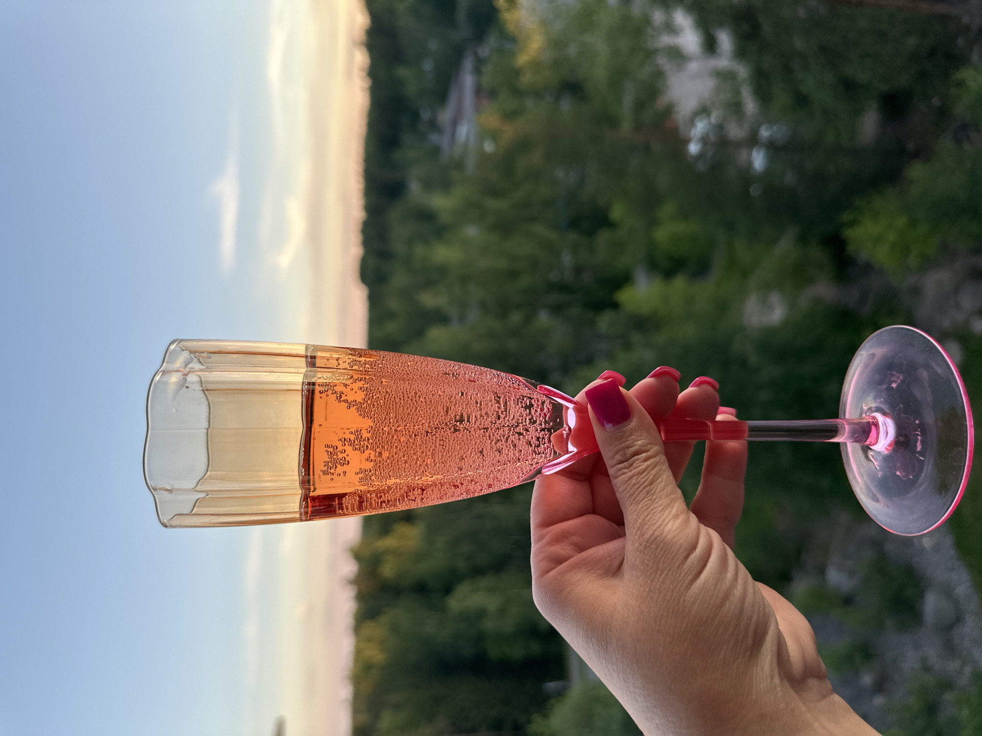 A close-up, eye-level shot shows a person's left hand, with bright pink fingernails, holding a champagne flute filled with a bubbly pink beverage. The hand is positioned in the lower left of the frame, with the fingers wrapped around the stem of the glass. The flute is a tall, slender shape with a ruffled rim and a pink stem and base, and it is filled to about three-quarters capacity. The drink inside is effervescent, with numerous small bubbles rising to the surface. The background, out of focus, depicts a lush, green forest canopy under a soft, dusky sky with hints of orange and pink from what appears to be a sunset. The overall scene suggests a moment of relaxation and enjoyment outdoors, possibly on a balcony or overlooking a natural landscape.