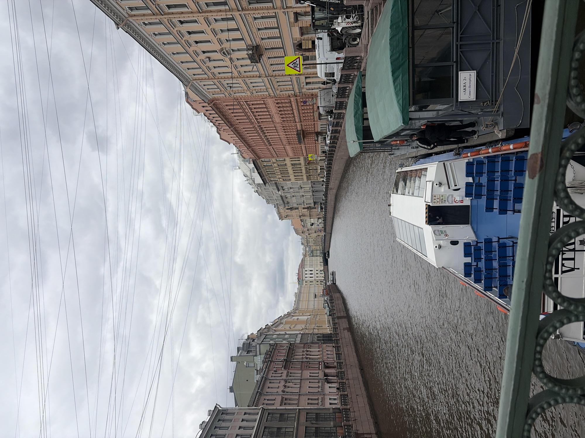 The image captures an urban canal scene in Saint Petersburg, Russia, viewed from a bridge on an overcast day. The narrow canal flows between historic buildings lining both banks.
On the left side, multi-story buildings display classical architecture in varying pastel shades of beige, pink, and yellow, adorned with decorative cornices and numerous windows.
The right bank features a paved embankment bordered by a green metal railing. Along this street, several vehicles, including vans and trucks, are parked. Visible signs include two yellow diamond-shaped warning signs with black symbols, and text "ЭЛЕКТРОМОНТАЖ" on one vehicle. A green-tarped structure is docked, bearing a white rectangular sign with black Cyrillic text: "АКВА 2ю" and "Санкт-Петербург". A person dressed in dark clothing stands on the dock beside this structure, looking towards the canal.
In the brownish canal water, a white and blue passenger boat is prominent. It has an open upper deck fitted with rows of blue seats and orange safety rings along its hull. A partial blue text, possibly "ПТА", is visible on its side.
The sky above is grey and cloudy, and numerous thin wires crisscross overhead, likely part of the city's infrastructure. In the immediate foreground, a decorative green metal railing with circular patterns, part of the bridge from which the photo was taken, is visible. The scene appears calm, depicting a typical day in the city with canal boat activity.