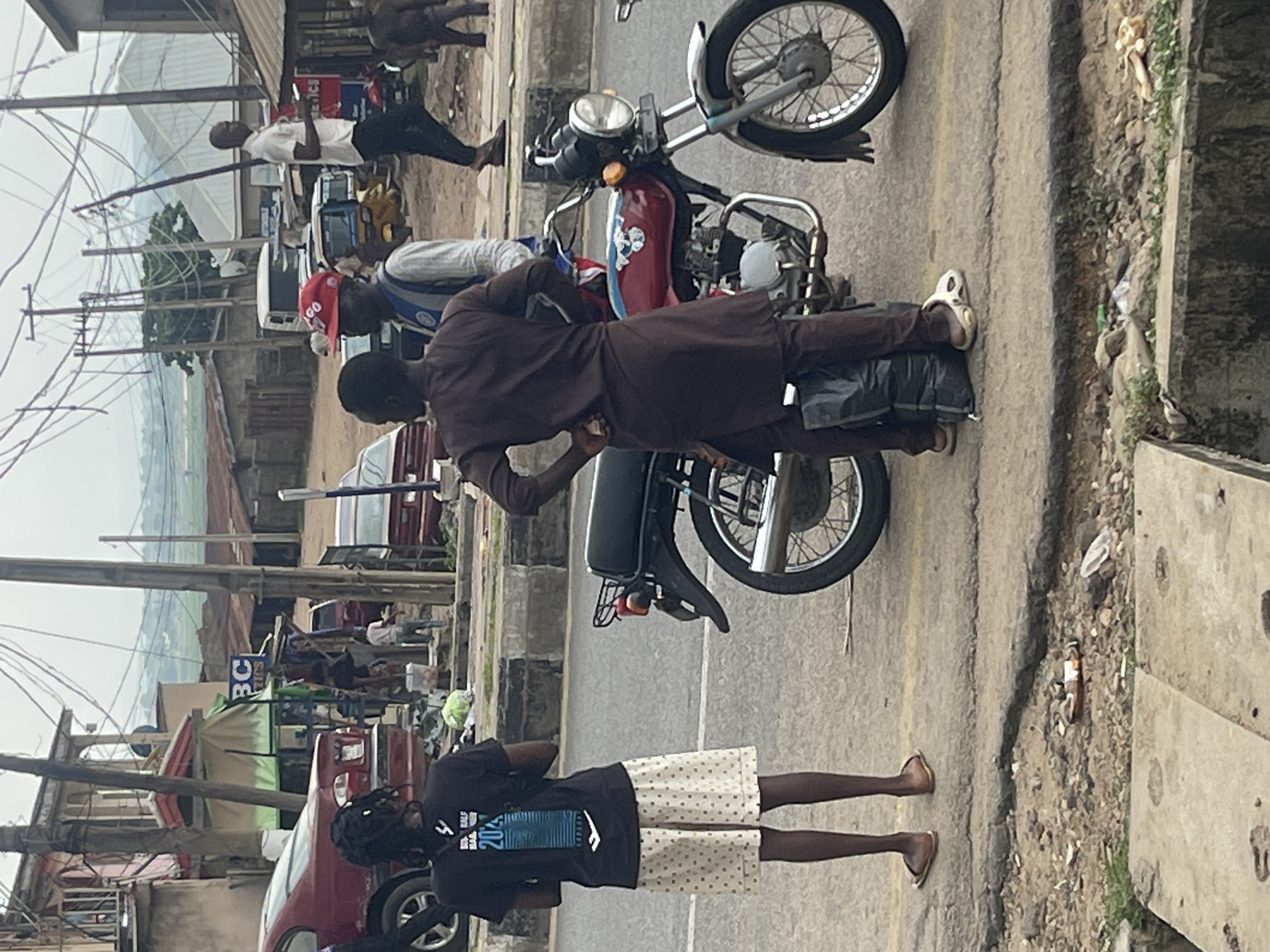 In the city of Unknown, Unknown, a street scene unfolds with several individuals and a motorcycle. A young woman with braided hair stands facing away from the camera, wearing a black t-shirt with a graphic design and a patterned skirt. Beside her, a person in a brown traditional garment with a sash stands with hands on hips, next to a black plastic bag. They are positioned near a red motorcycle with a blue stripe and a white emblem on the fuel tank, which has a chrome exhaust pipe and a luggage rack. Another man in a white collared shirt and dark pants is visible in the background, walking away from the motorcycle. The environment is characterized by overhead wires, utility poles, and buildings with tin roofs, suggesting an urban or semi-urban setting. The ground is paved with some debris and a roadside ditch.