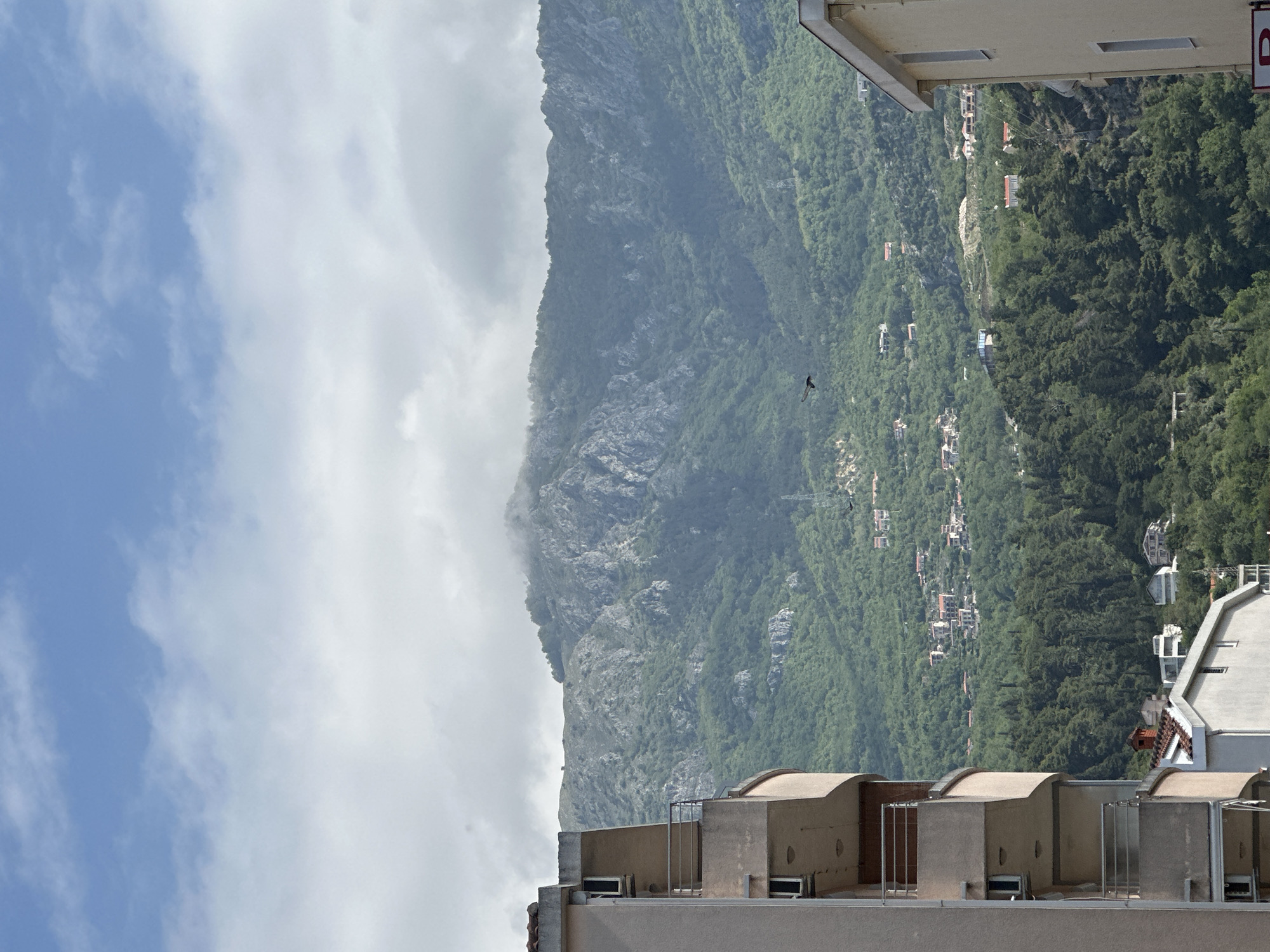 The image depicts a mountainside landscape adjacent to an urban area, likely in Budva, Montenegro. The left side of the frame features a sky with patchy white clouds against a light blue background. A prominent, steep mountain face ascends vertically, covered in patches of grey rock and dense green vegetation. Dotted along the lower slopes of the mountain are residential buildings, suggesting a settlement in the foothills. The right side of the frame shows sections of buildings, appearing as if the image was taken from an elevated position, possibly a rooftop. The architecture is modern, with rounded structures and a muted color palette. A small bird is visible in flight against the mountain backdrop.